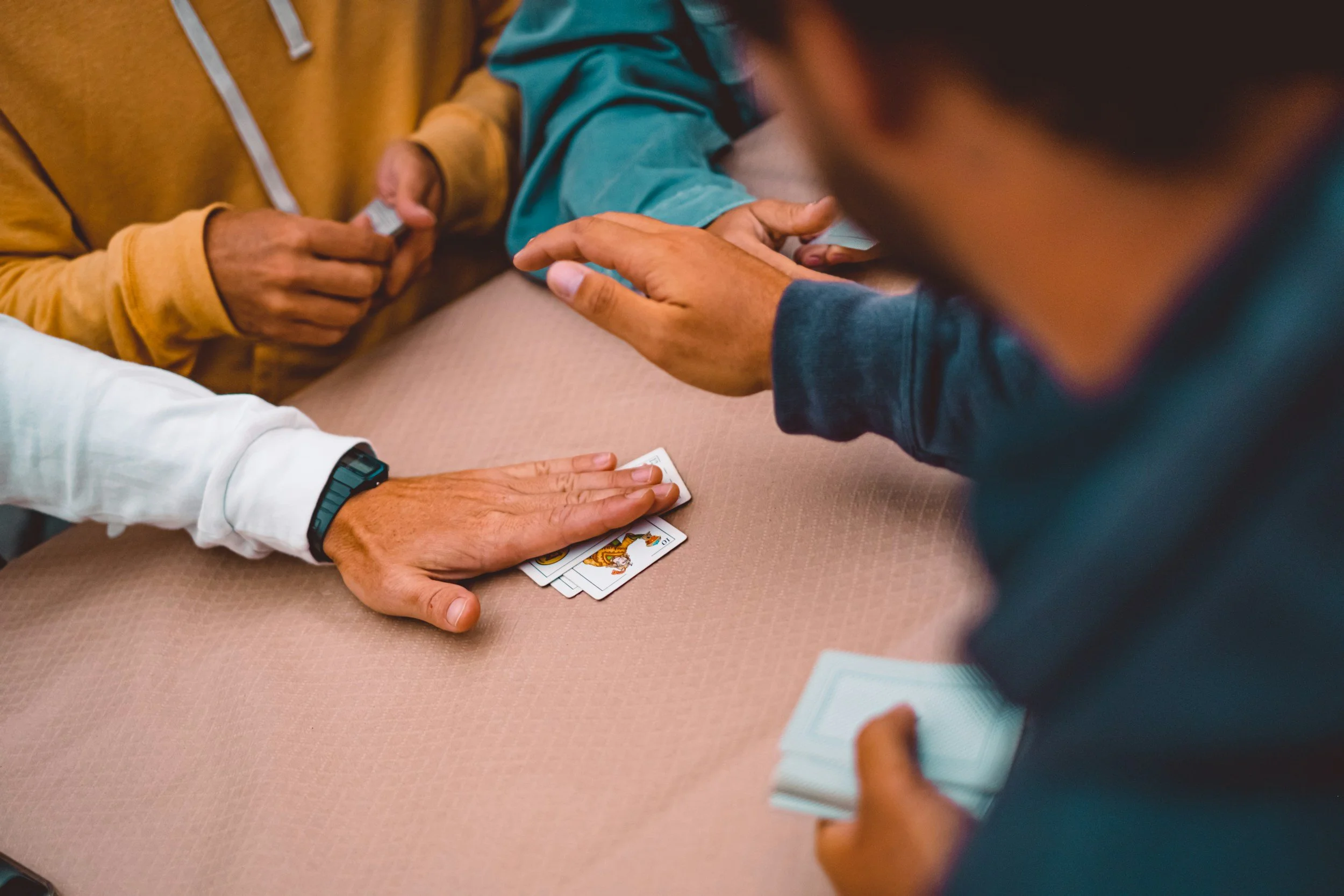 Close-up of a deck of playing cards on a table, with a hand holding the deck, and another person placing a card on the table. The focus is on the hands and cards, with other people's bodies in the background.