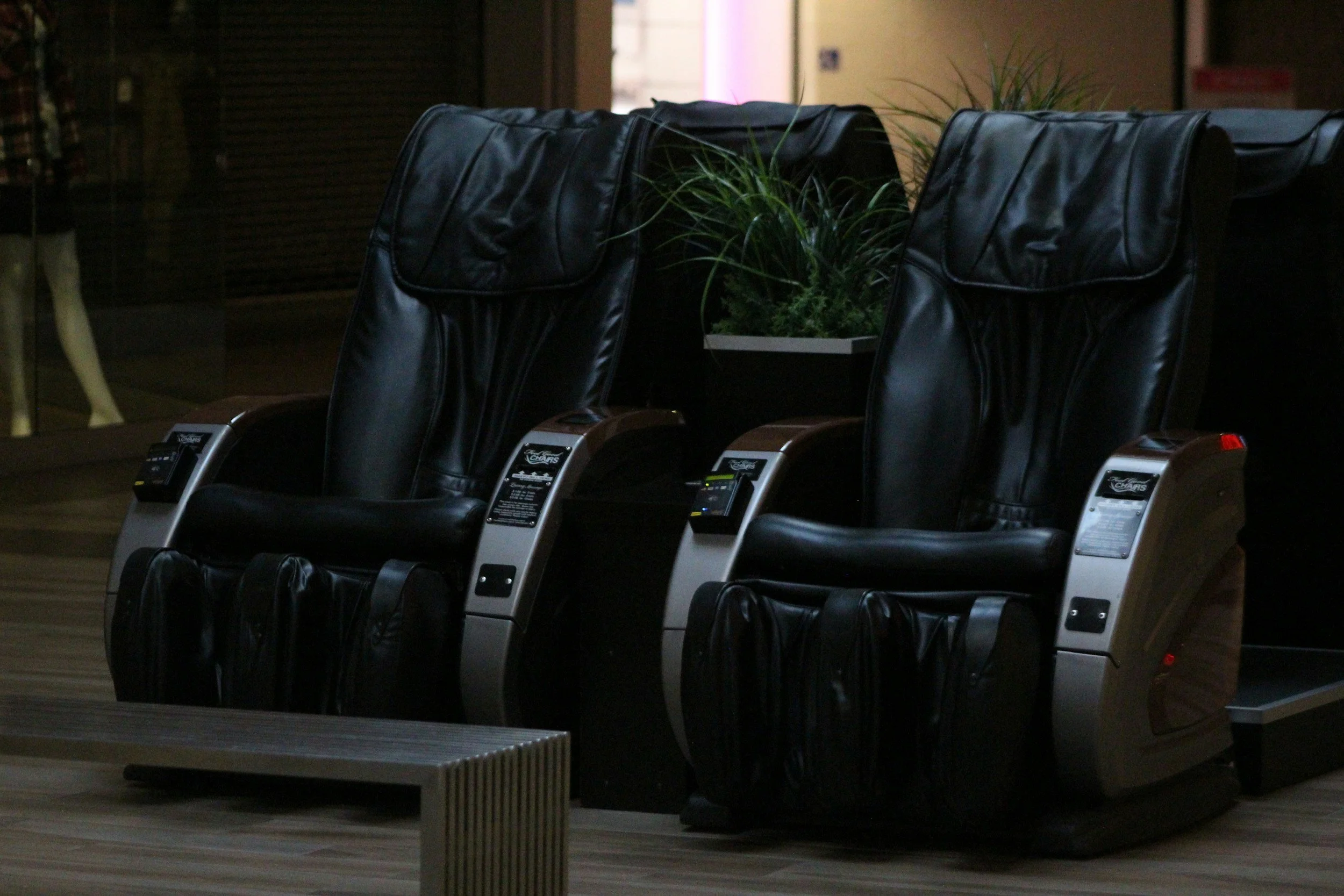 Row of massage chairs with black leather upholstery and control panels, positioned along a wooden floor with a potted plant in the background.