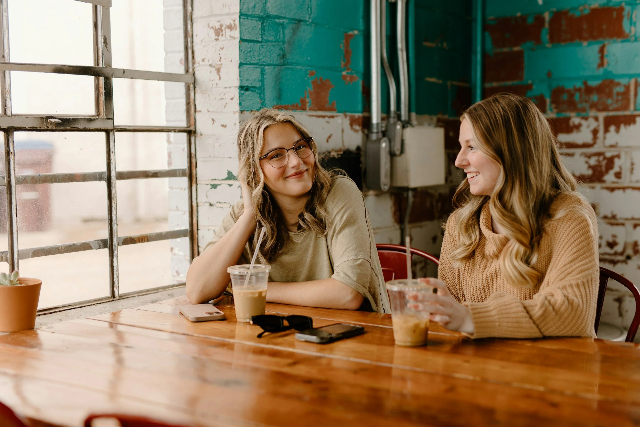 Two women sitting at a wooden table, smiling and chatting, with iced coffee drinks in front of them, in a rustic cafe with exposed brick and teal painted walls.