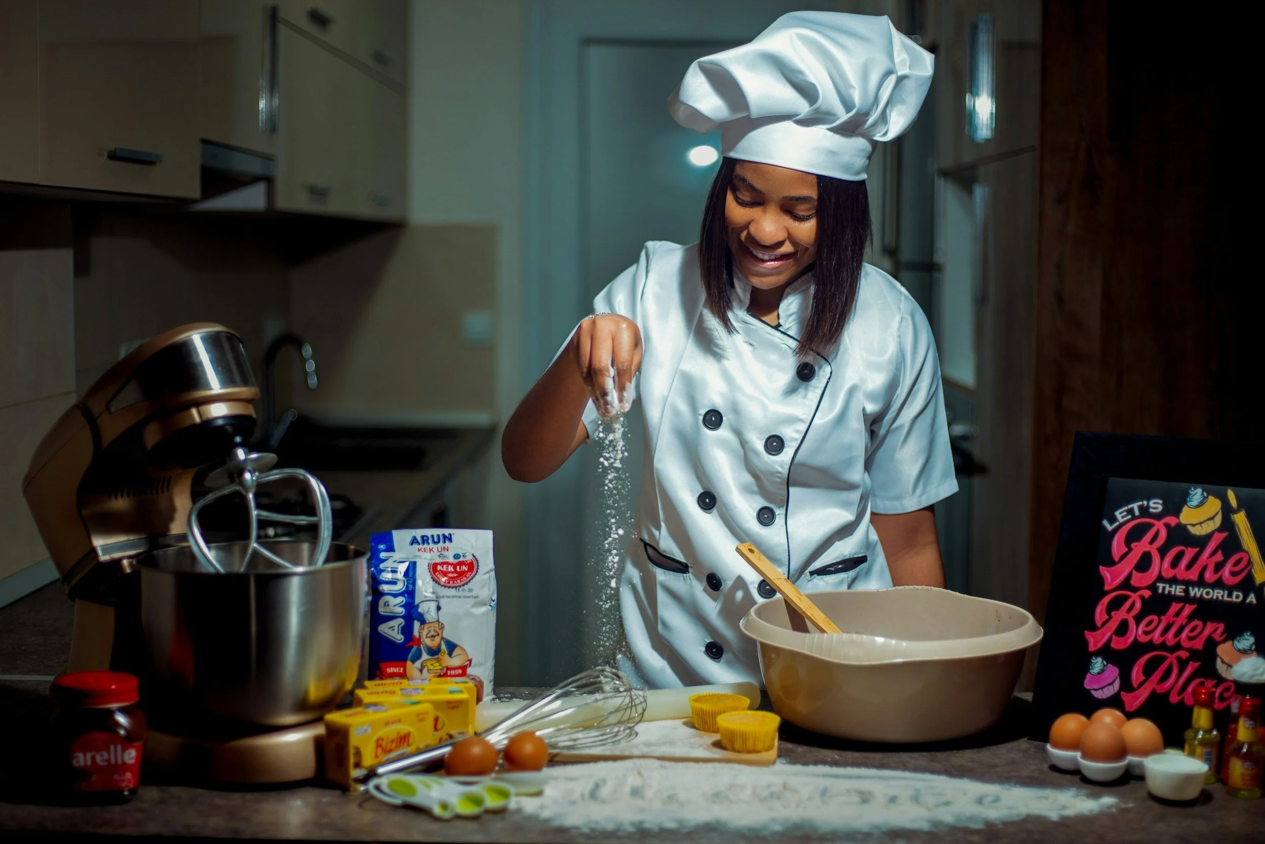 A female chef in a white uniform and chef hat is smiling while sprinkling flour on a table in a kitchen. Various baking ingredients and tools are spread across the countertop, including eggs, butter, measuring cups, and a mixer.