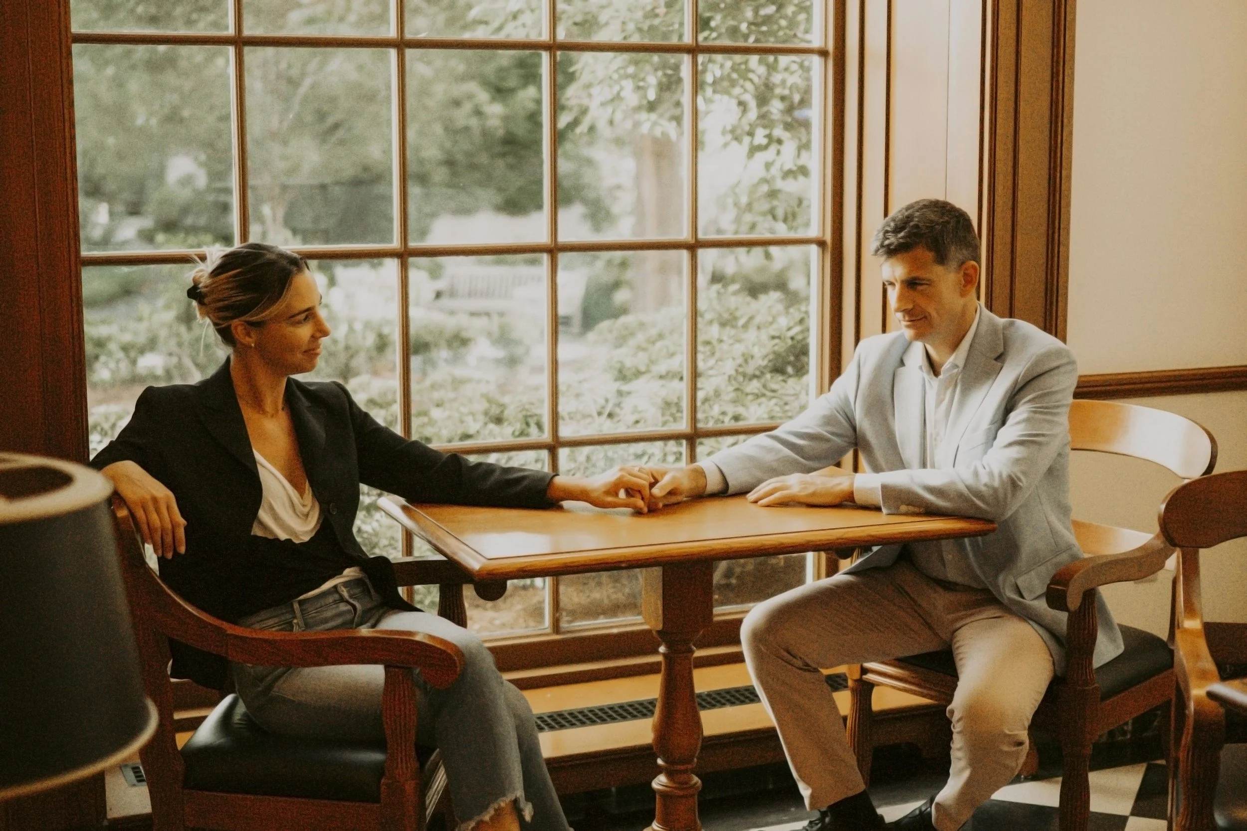 A man and woman sitting at a table in a room with large window panes, holding hands across the table, facing each other and smiling, with lush greenery visible outside.