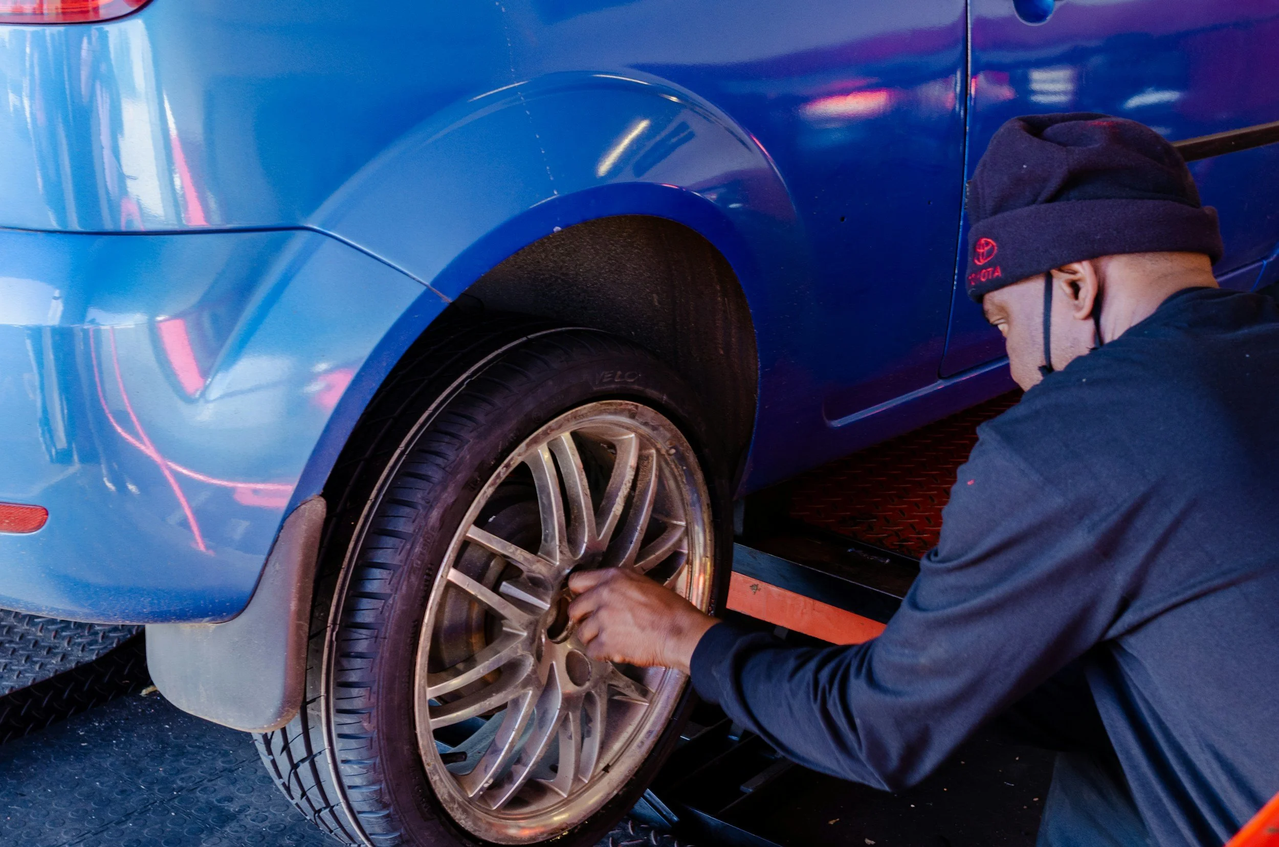 A person wearing a black hoodie and a beanie with a Toyota logo is working on a blue car, specifically on the front left wheel.