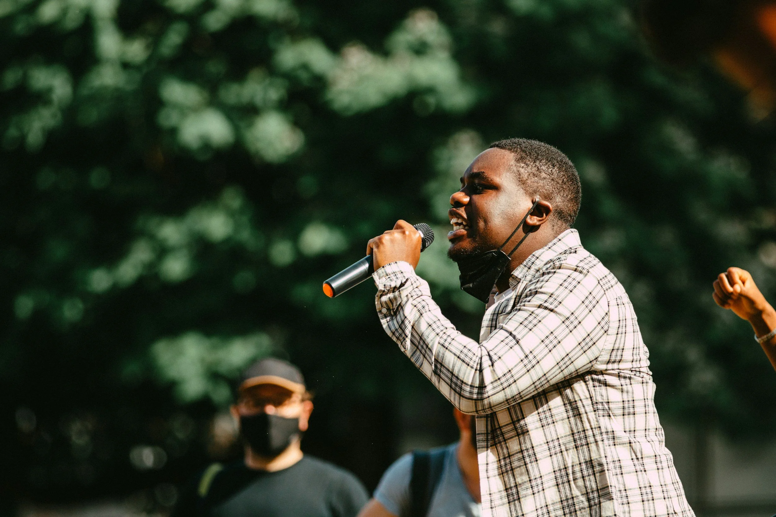 A man wearing a plaid shirt and a face mask, speaking into a microphone during an outdoor event with others in the background against a backdrop of green trees.