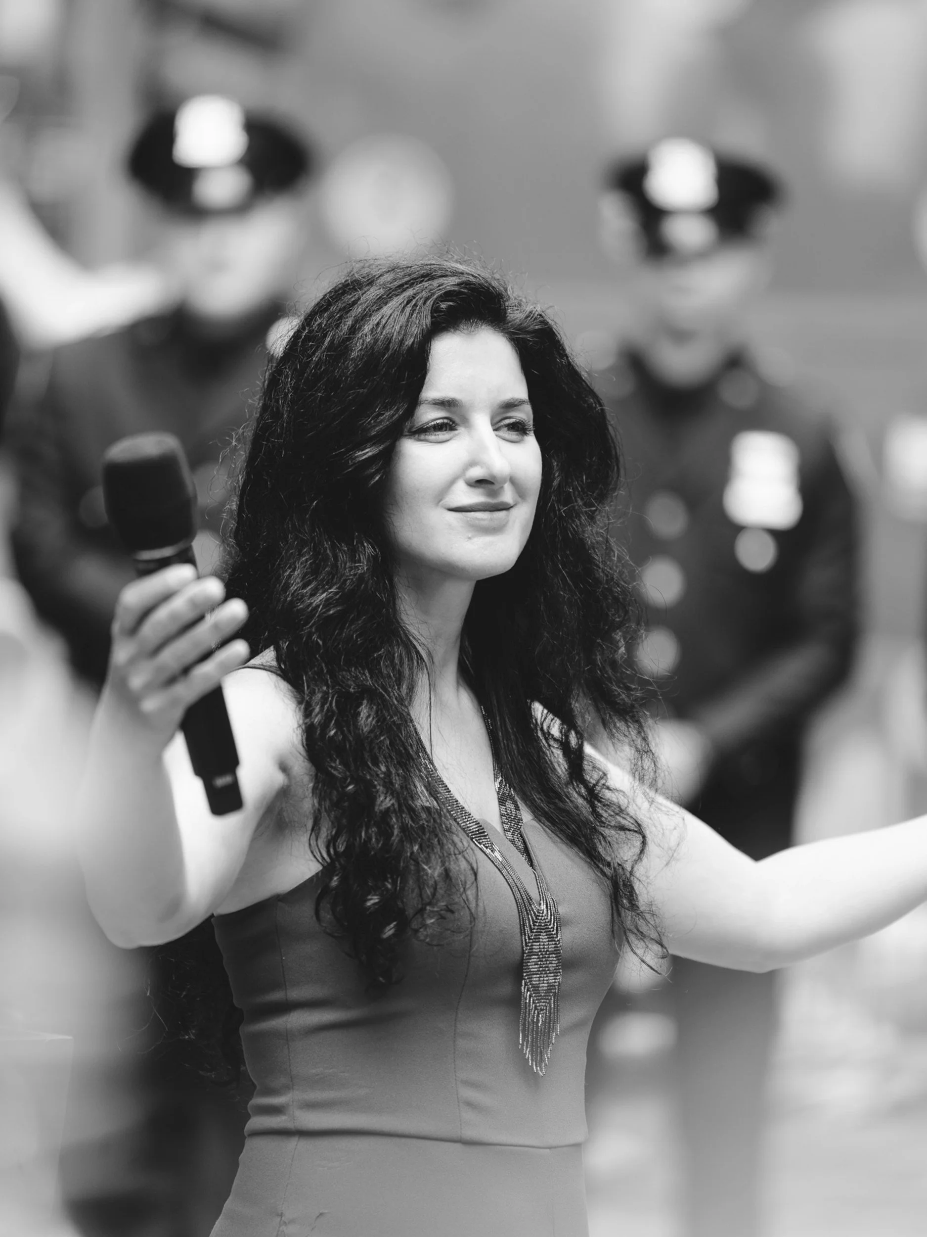 Black and white photo of a woman with long, wavy hair holding a microphone in her right hand and smiling, with two young men blurred in the background.