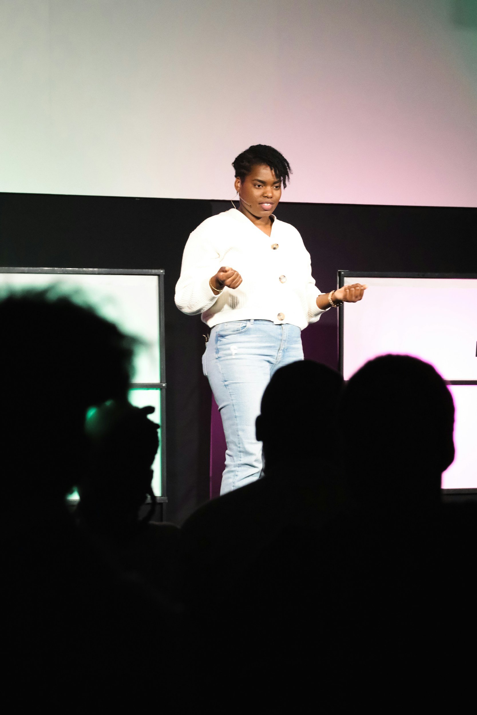 A woman with short curly hair standing on stage with a large screen behind her, speaking or presenting to an audience.