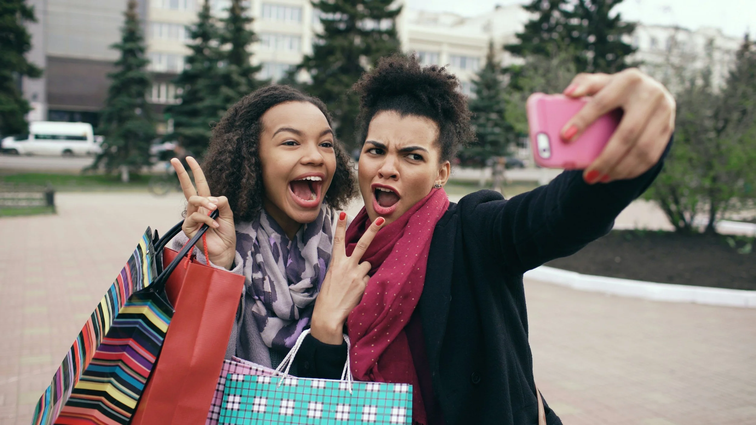 Two women taking a selfie outdoors, both making playful facial expressions and peace signs. One woman has curly hair and is holding shopping bags, while the other has natural hair and is holding a pink phone.