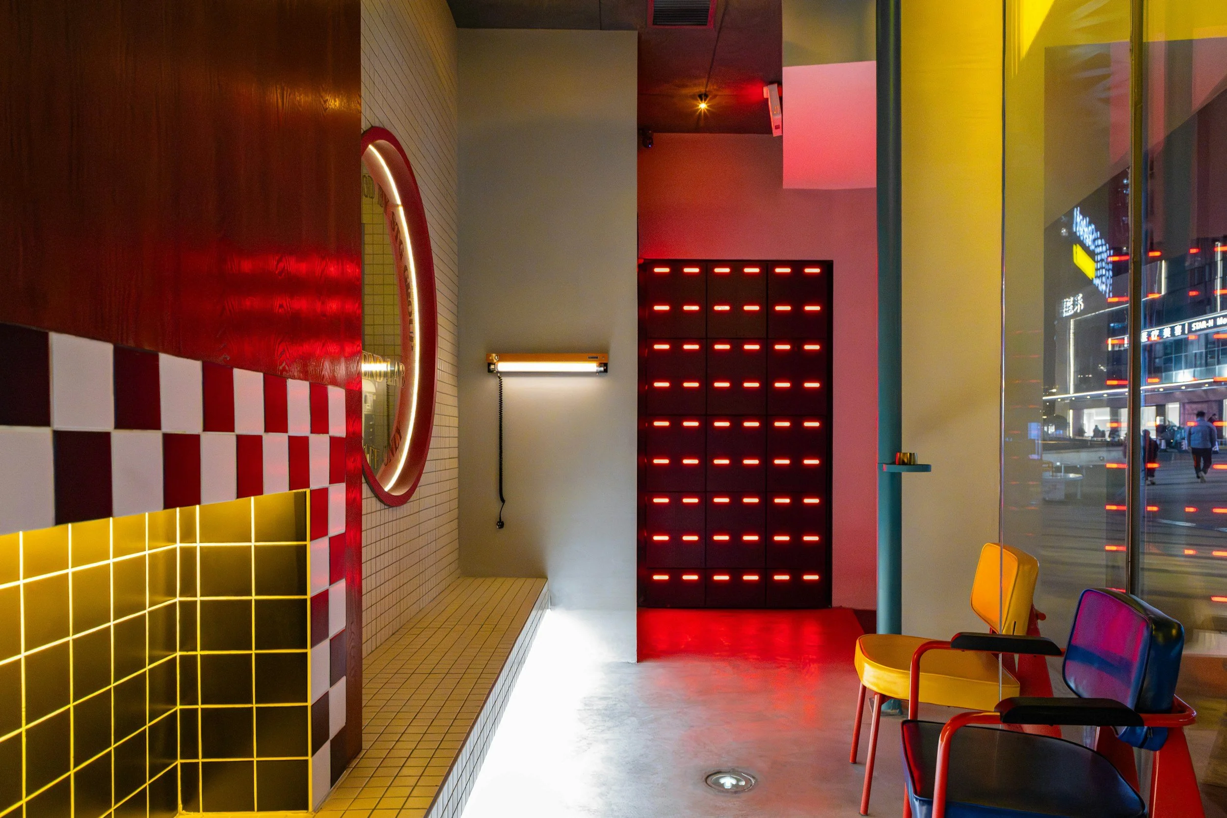 Interior of a modern fast-food restaurant with colorful chairs, a tiled counter with a yellow and black checkered pattern, red and black illuminated decor, and large windows showing a city street at night.