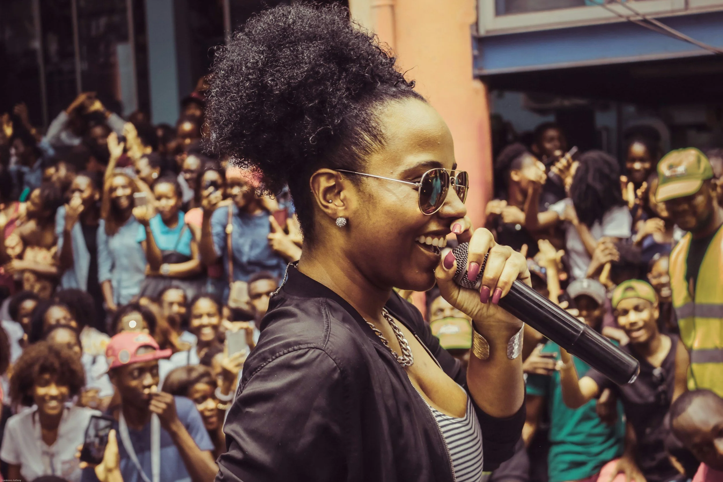 A woman with dark curly hair in a high puff, wearing sunglasses, earrings, and a black leather jacket, smiles and holds a microphone. She is surrounded by a crowd of people, mostly women, in an outdoor setting.