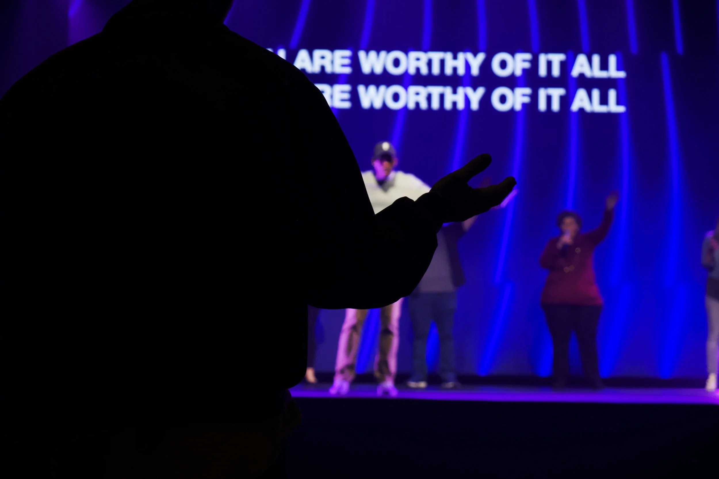 A silhouette of a person pointing towards a stage with performers and a large illuminated screen with the words "You are worthy of it all" in the background.