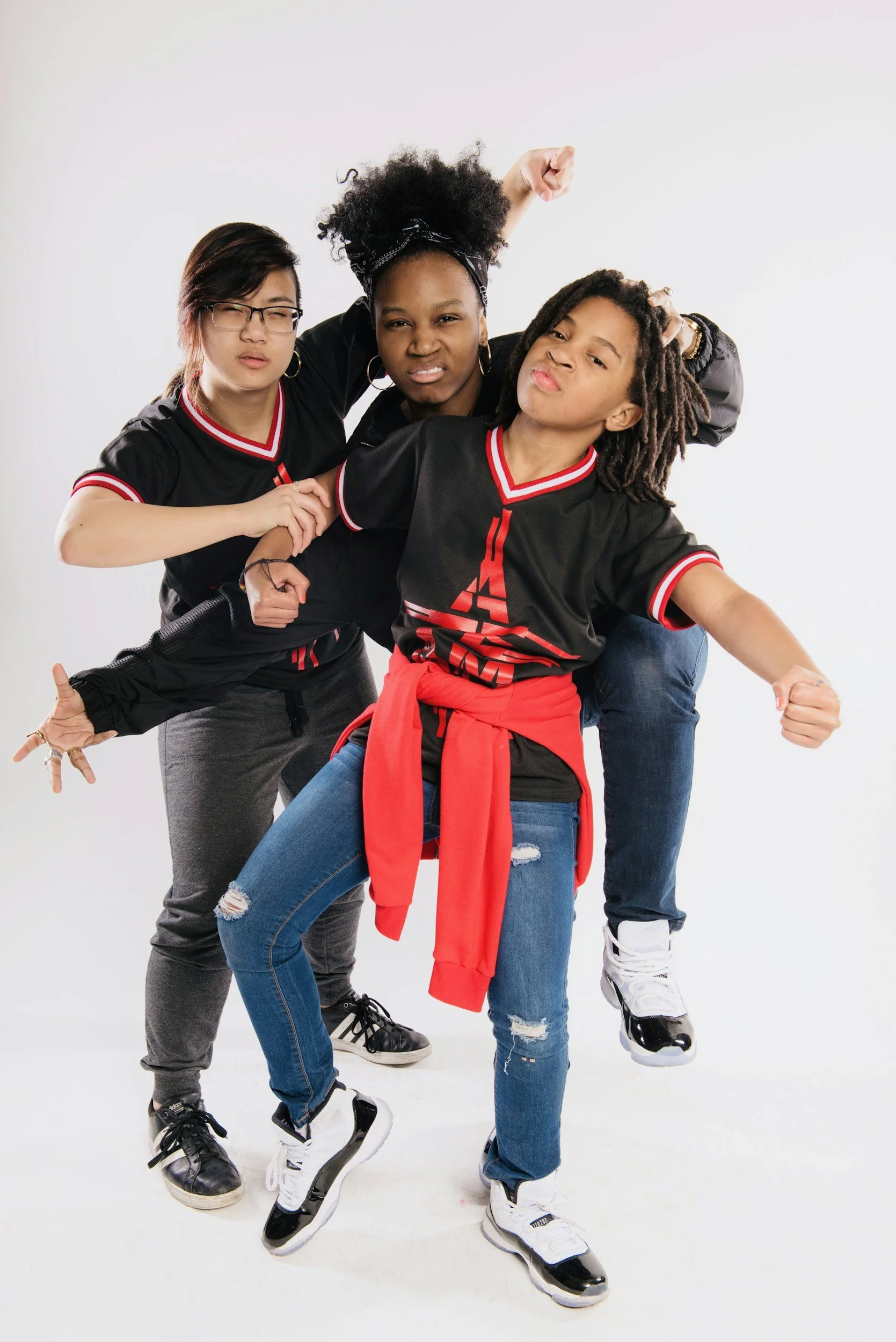 Four young women posing together in casual clothes, with one kicking up her leg, in front of a white background.