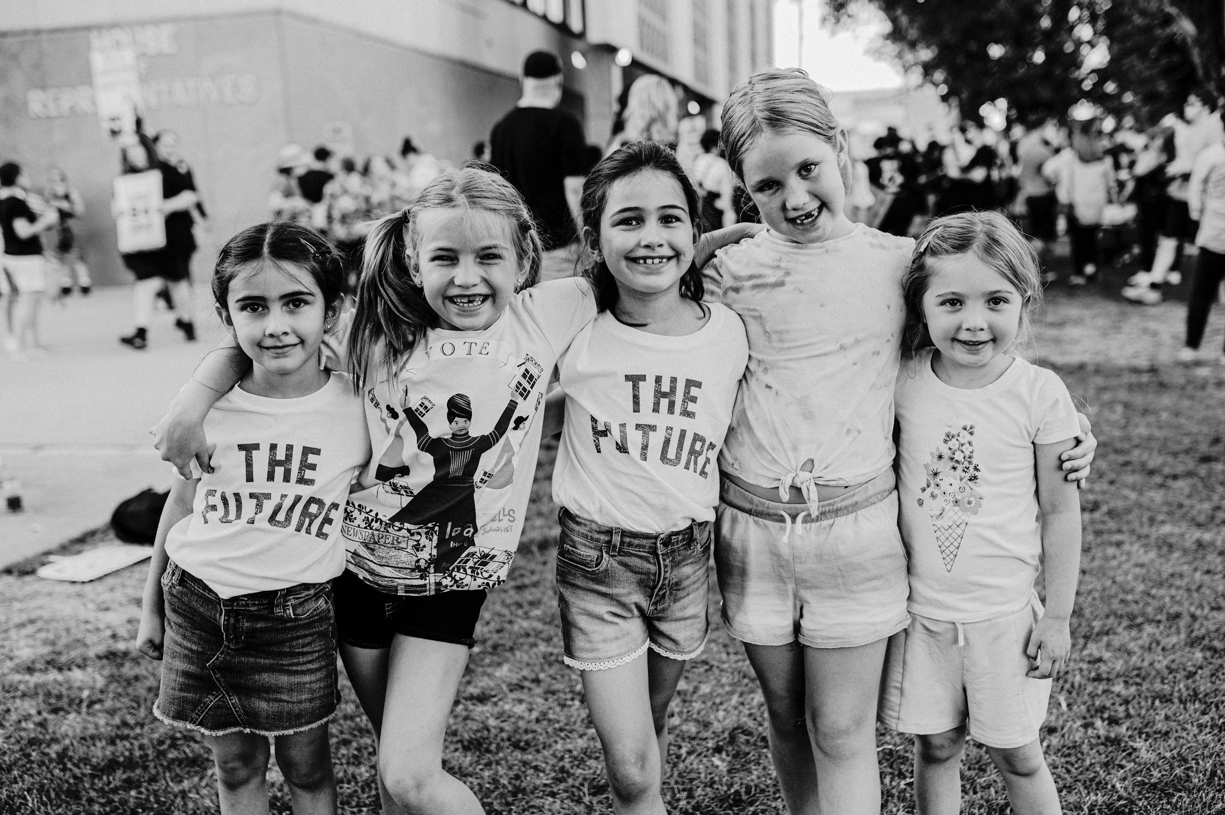Group of five young girls smiling with their arms around each other at an outdoor event, with other people blurred in the background.
