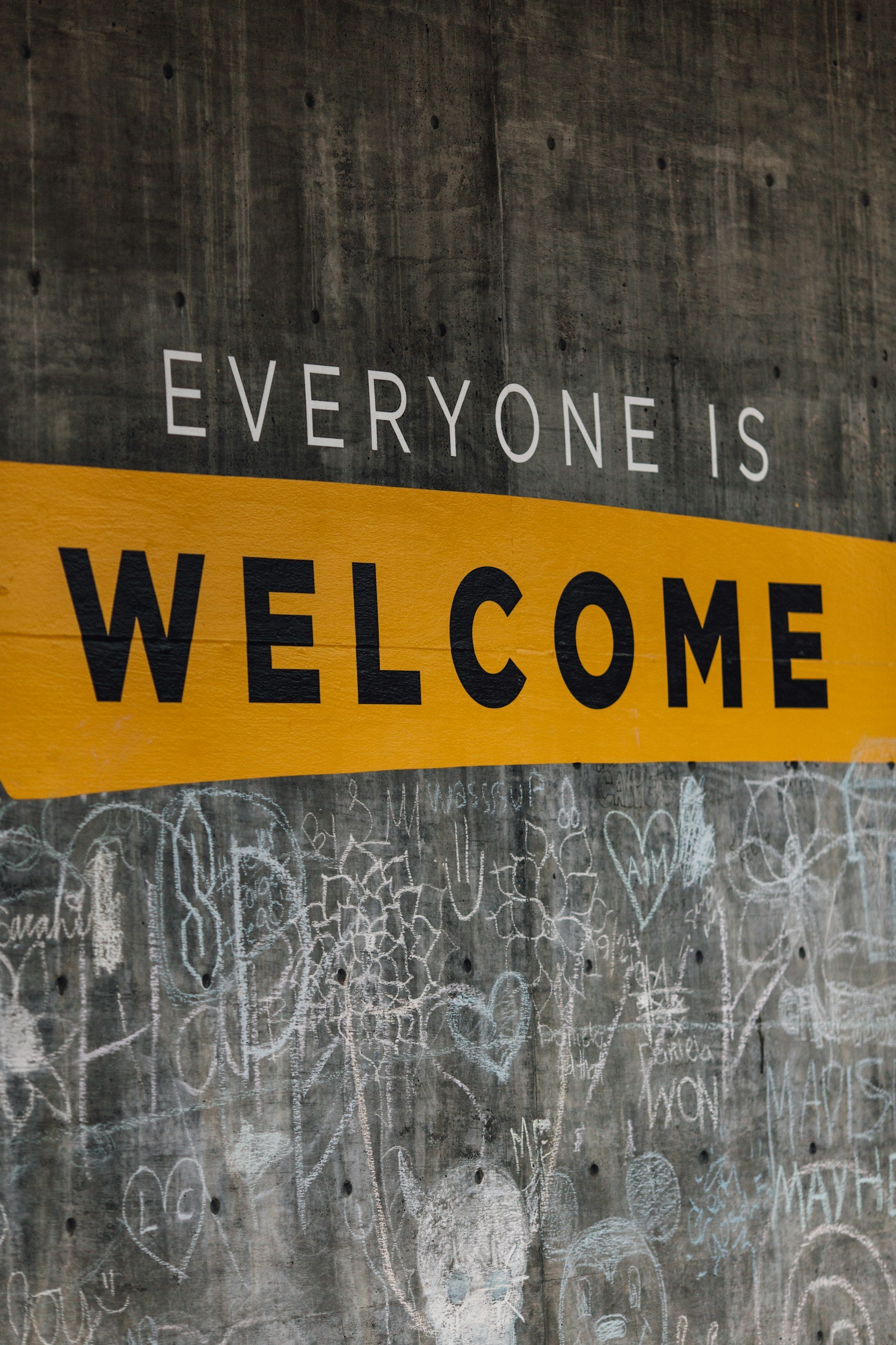 A concrete wall with a yellow and black striped banner that reads, 'Everyone is Welcome.' The wall is covered in chalk drawings and writings, including hearts and various phrases.