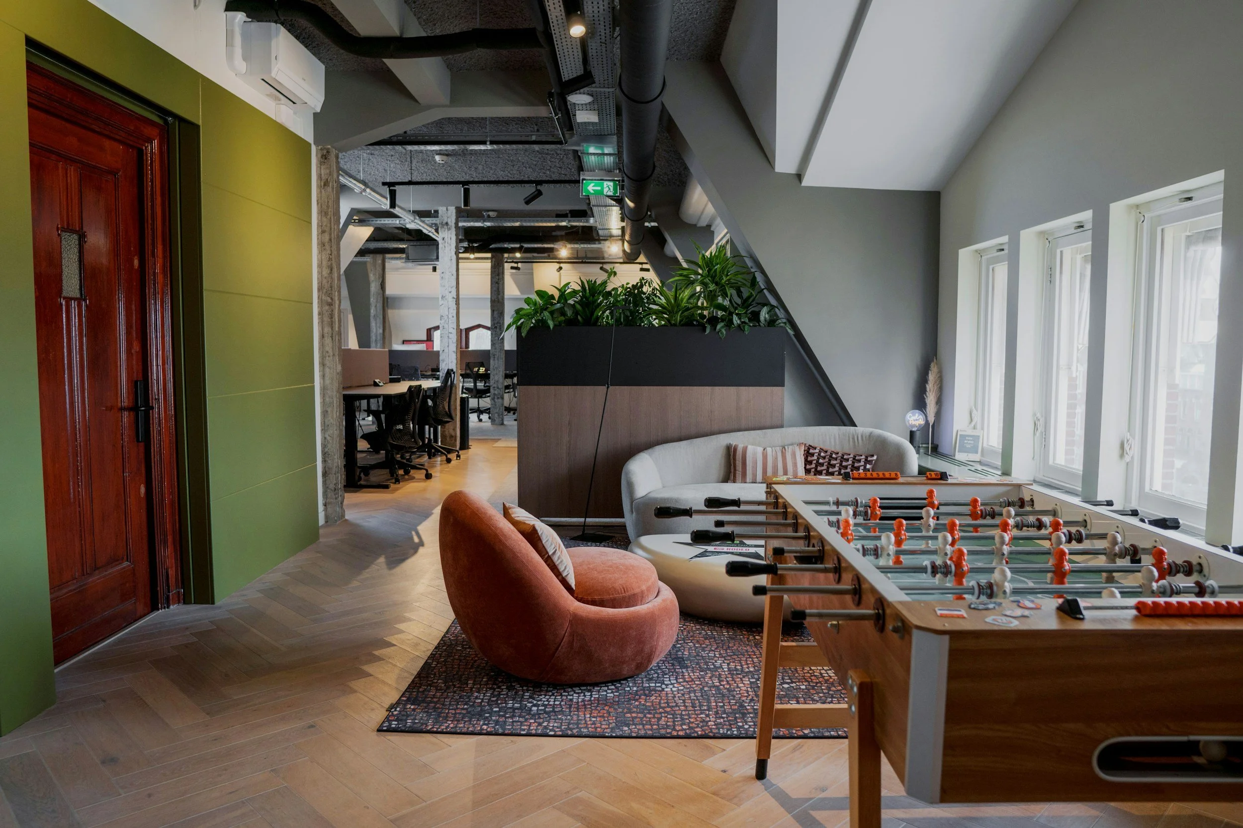 Modern office space with a foosball table, a white sofa with striped pillows, and large windows letting in natural light. There's a green wall, indoor plants, and office desks in the background.