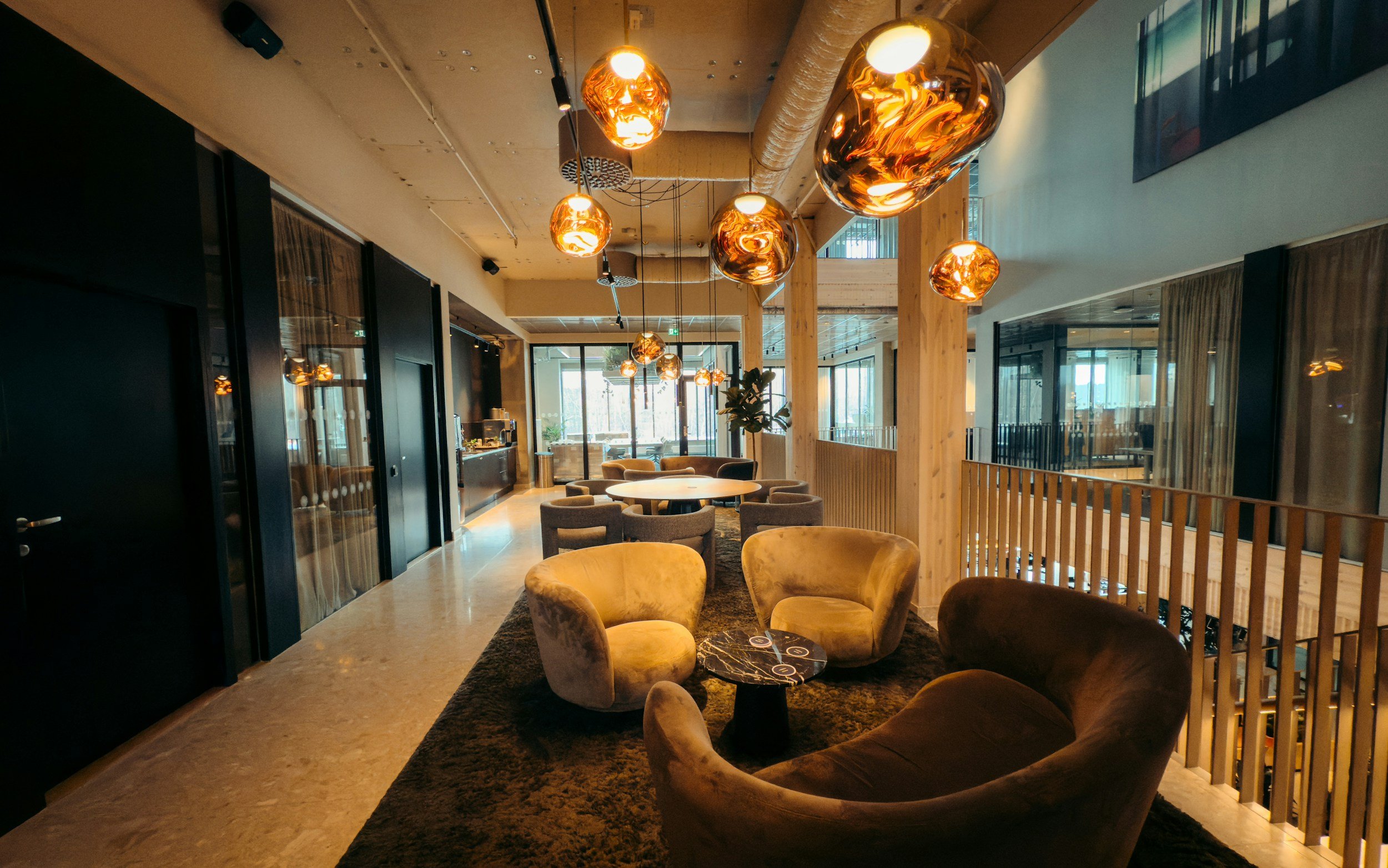 Interior of a modern lounge or cafe with round yellow armchairs, a black marble table, a dark rug, and unique orange glass pendant lights hanging from the ceiling. There are large windows and glass doors allowing natural light, with wooden beams and black walls.