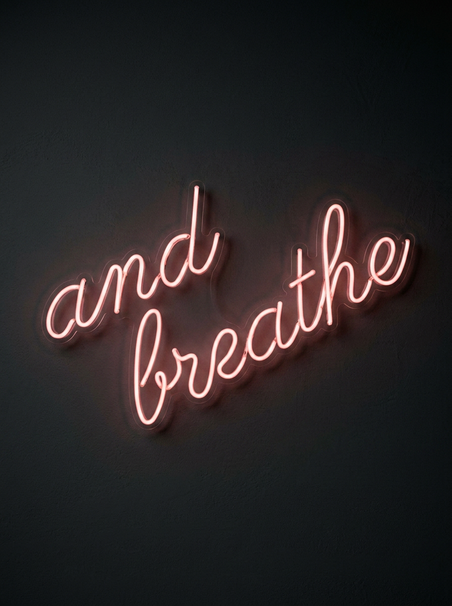 Pink neon sign on a dark wall that reads 'and breathe' in cursive writing.