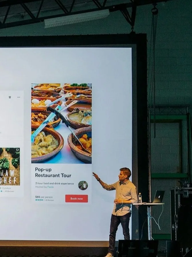 A man giving a presentation in a large industrial-style room with a huge projection screen displaying a food and drink experience tour advertisement. The screen shows images of bowls of food and tilting utensils. The presenter stands at a small table with a laptop and a water bottle.