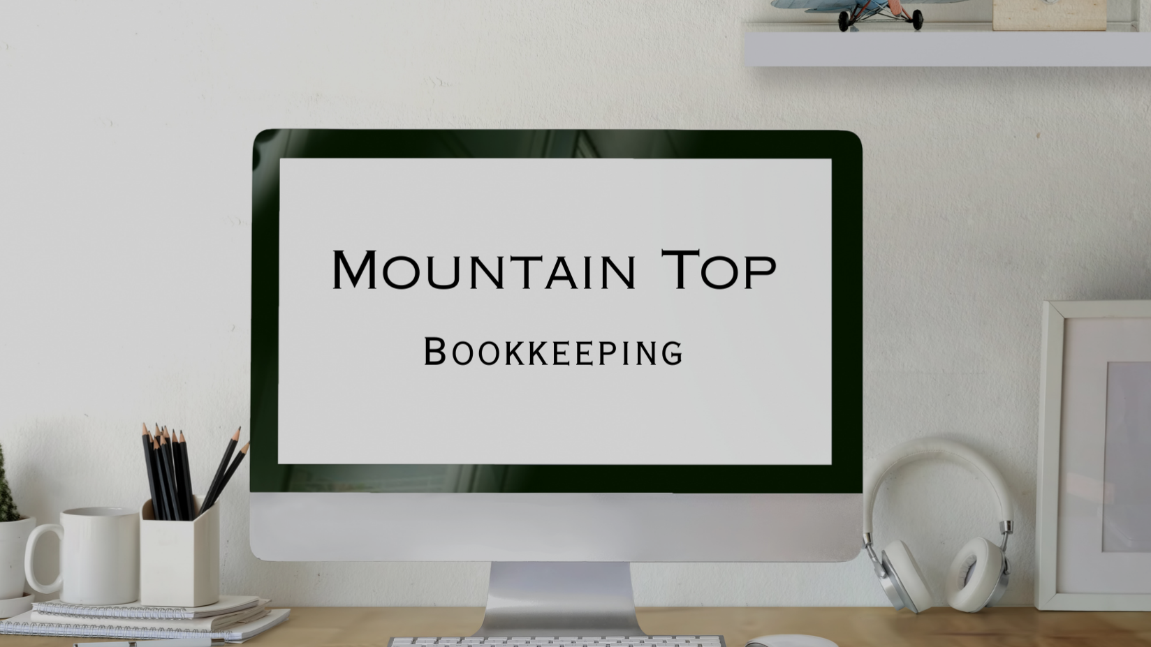 Computer screen displaying 'Mountain Top Bookkeeping' on a white background, workstation with black pencils in a white mug, white mug, notebook, and headphones on a wooden desk.
