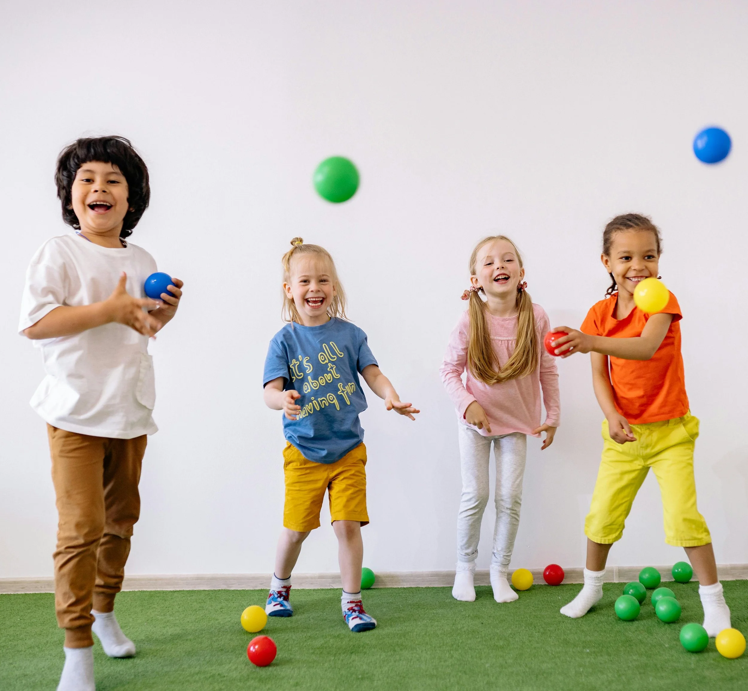Four children playing with colorful balls in an indoor setting with a grassy floor and plain white wall.