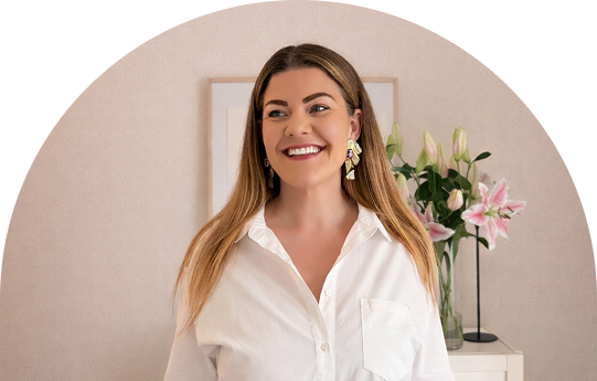 A woman smiling in front of a pink and black background, with pink lilies on a side table behind her.