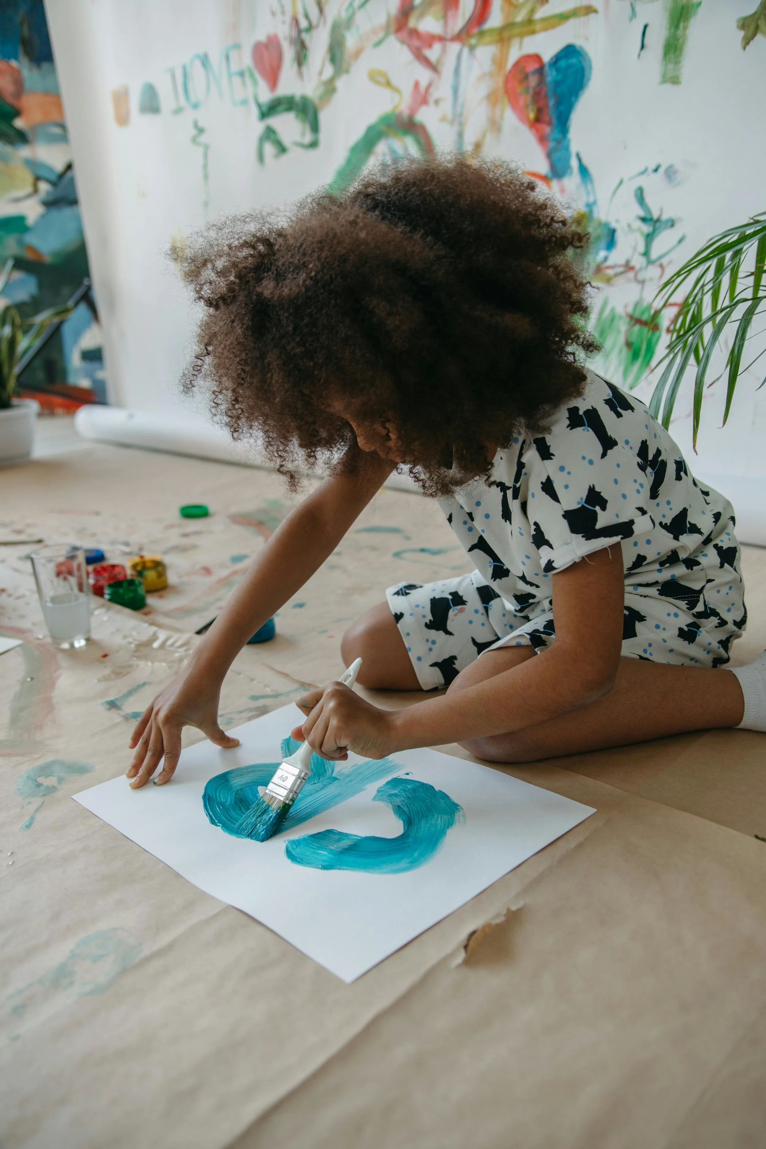 A young girl with curly hair is kneeling on the floor and painting with a brush on a sheet of white paper. She is wearing a pajamas with bear prints and is surrounded by art supplies, and the background features a colorful, childlike mural on the wall.