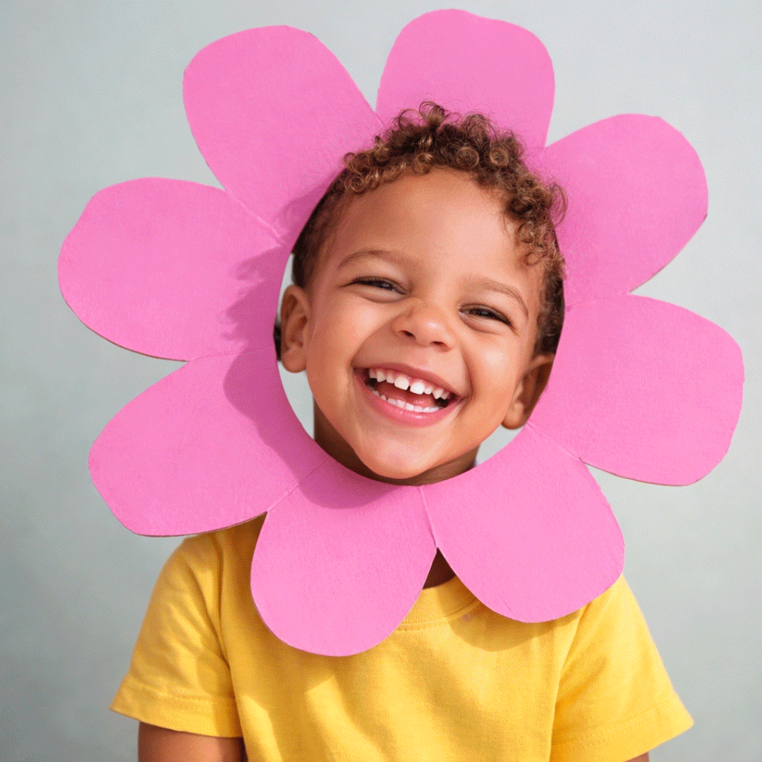 JOY! Childcare Nanny Agency in London, UKA young child with curly hair smiling wide, wearing a yellow shirt, behind a large pink flower cutout framing her face.