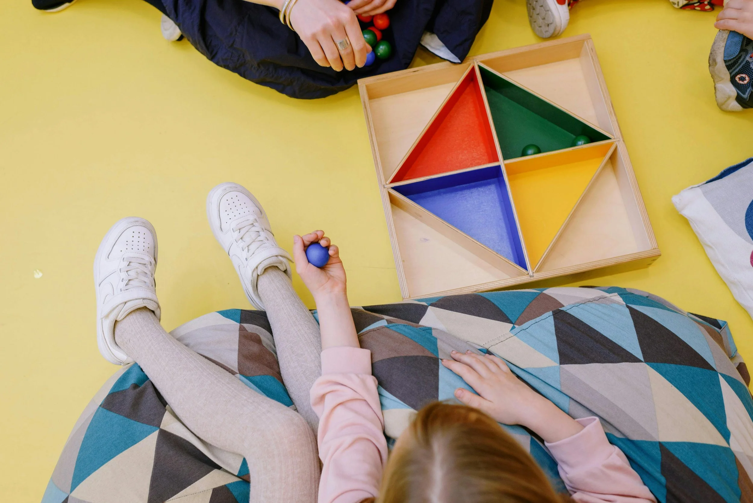 A child sitting on a colorful geometric-patterned cushion or blanket, holding a blue ball, with a wooden triangle stacking toy with colored sections and balls, and an adult's hands reaching towards the toy, on a yellow floor.