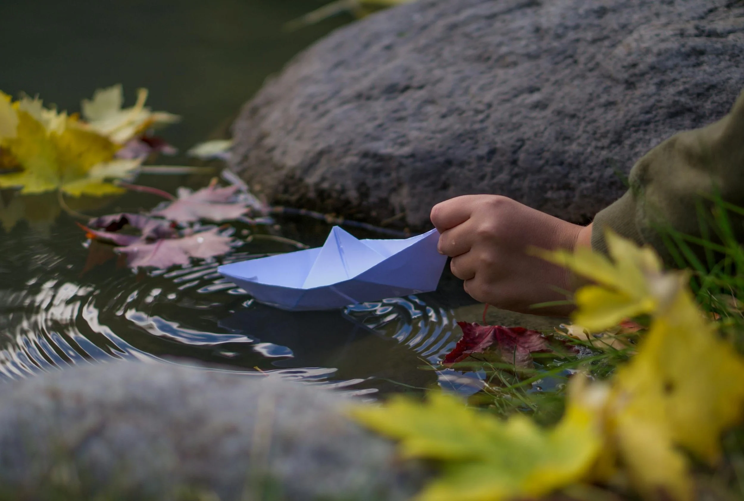 A person releasing a paper boat into a small pond surrounded by fallen autumn leaves and rocks.