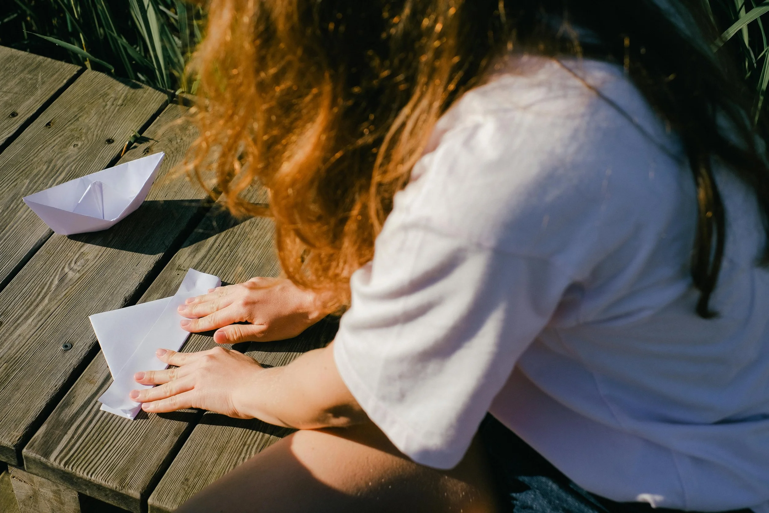 Person with long red hair sitting on a wooden dock, folding a paper boat, with another paper boat nearby in an outdoor setting.