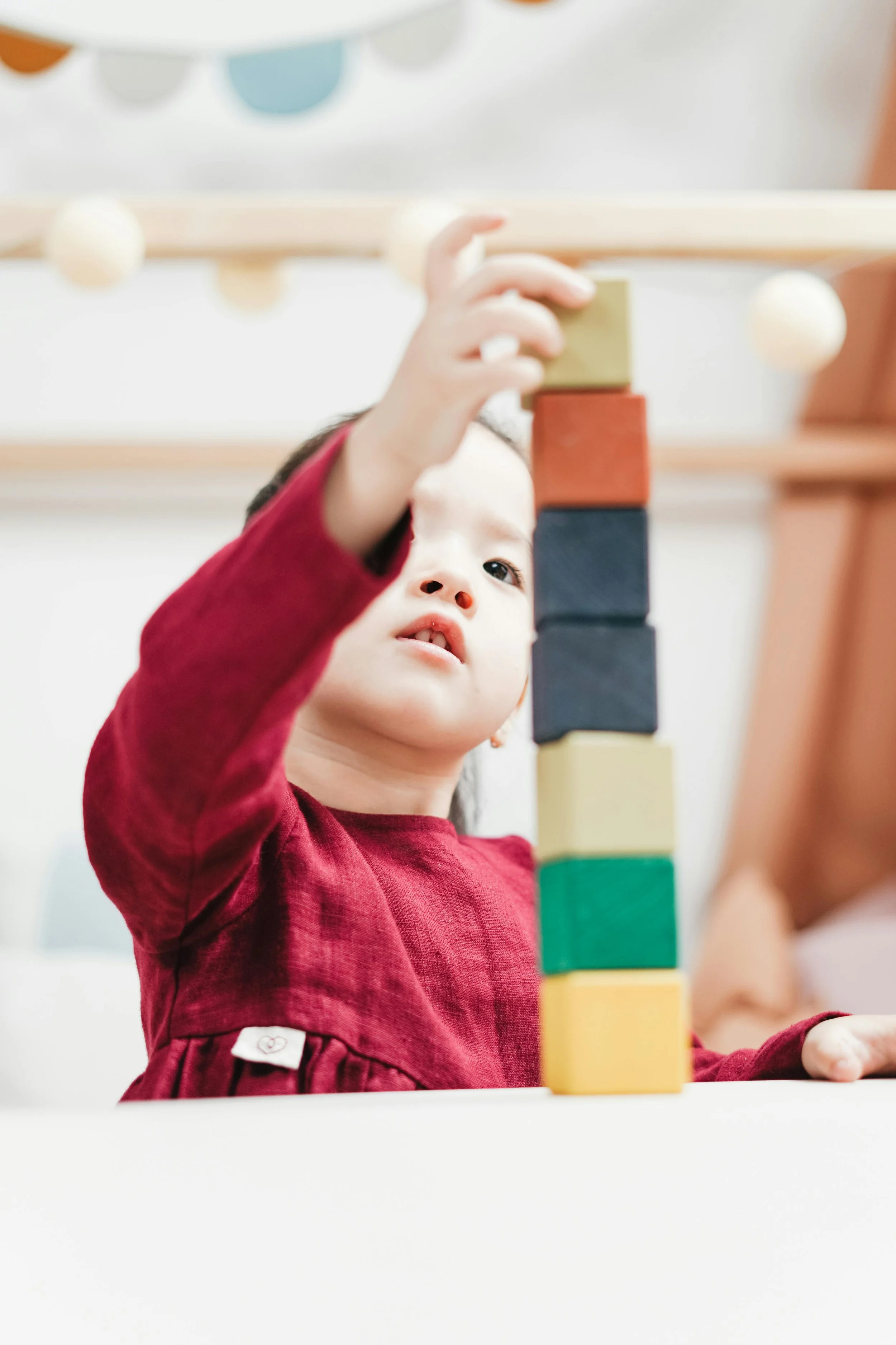Young girl stacking colorful wooden blocks, wearing a red dress, inside a playroom or classroom. JOY! Childcare Nanny Agency in London, UK