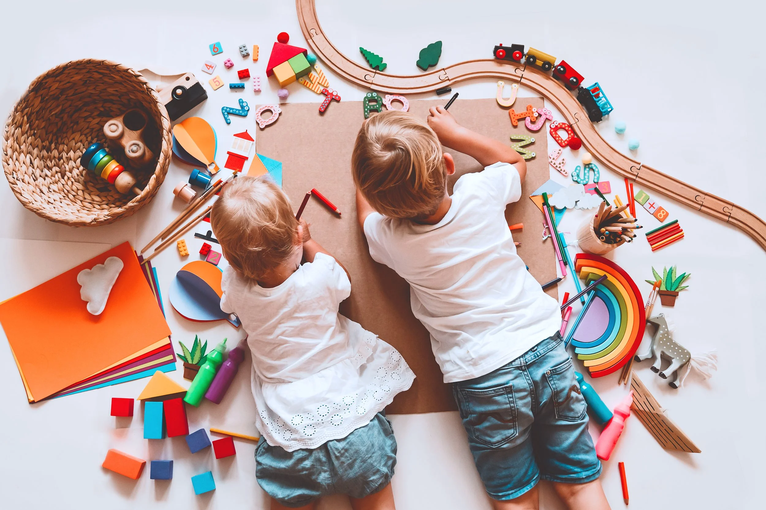 Two children playing with colorful toys, including train tracks, building blocks, and art supplies, on a white surface.
