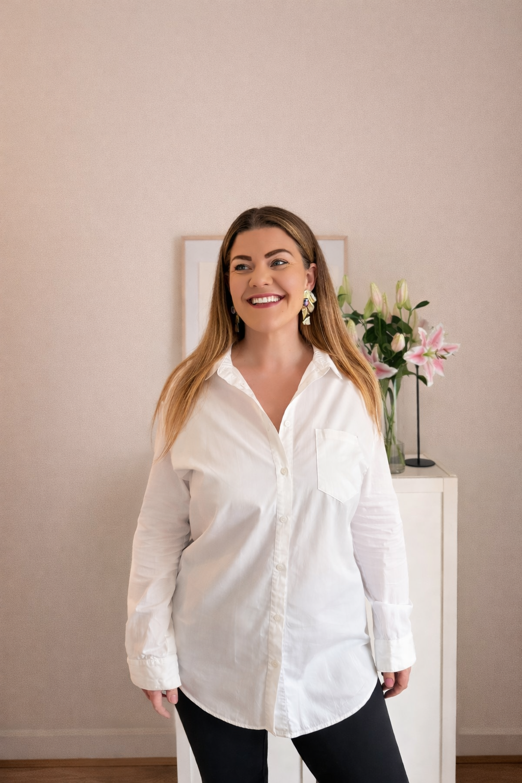 A smiling woman with long brown hair wearing a white button-up shirt and black pants, standing in a room with a beige wall, a framed picture, and a vase of pink lilies on a white cabinet behind her. JOY! Childcare Nanny Agency in London, UK