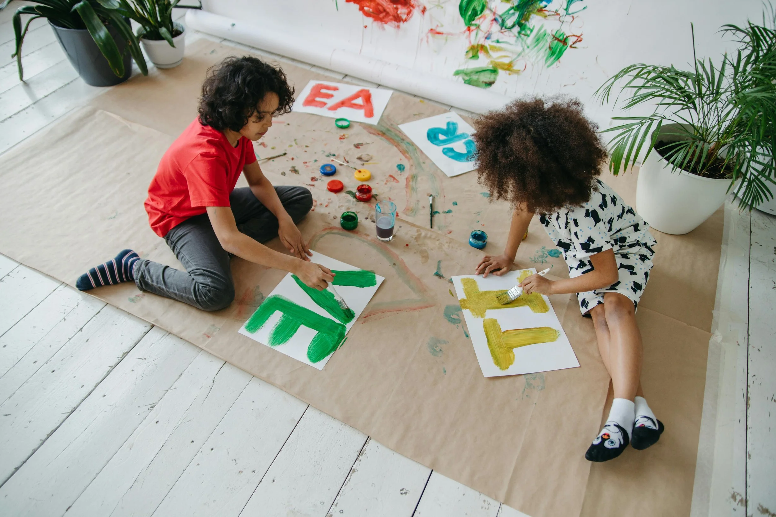 Two children painting on paper while sitting on a large sheet of brown paper spread on the floor, surrounded by colorful paint pots and brushes, with a wall decorated with painted letters in the background.