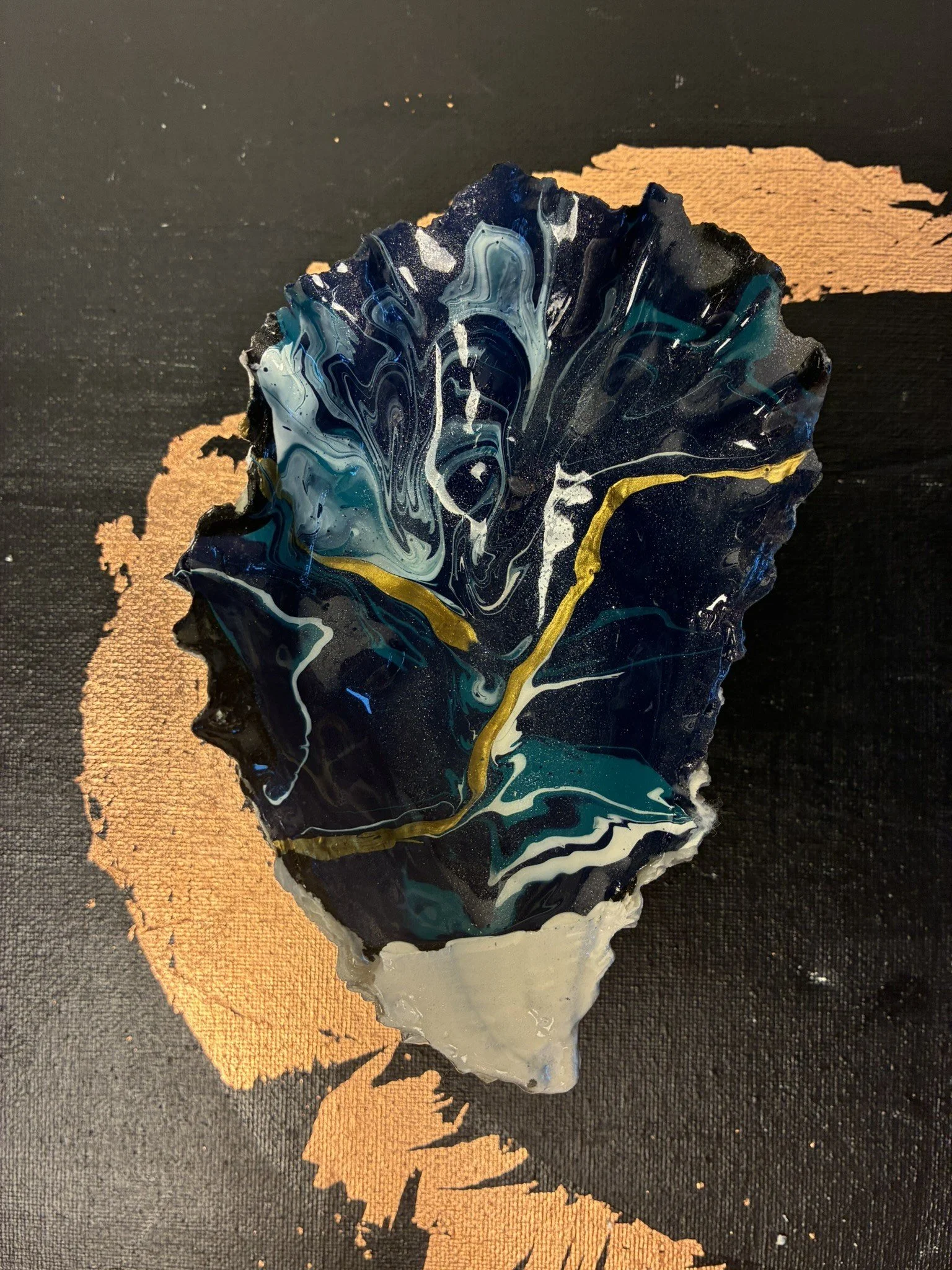 Abstract art piece featuring a large, one of one never replicated oyster shell with swirling blue, black, white, and gold colors, placed on a black surface with a copper leaf cresent.
