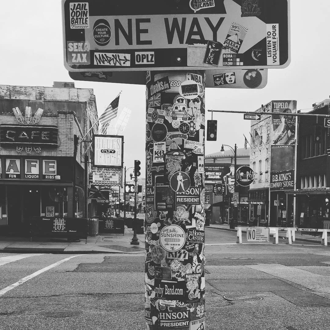 Street corner with a pole covered in various stickers, a 'ONE WAY' traffic sign above, and surrounding buildings with signs and awnings, in an urban area
