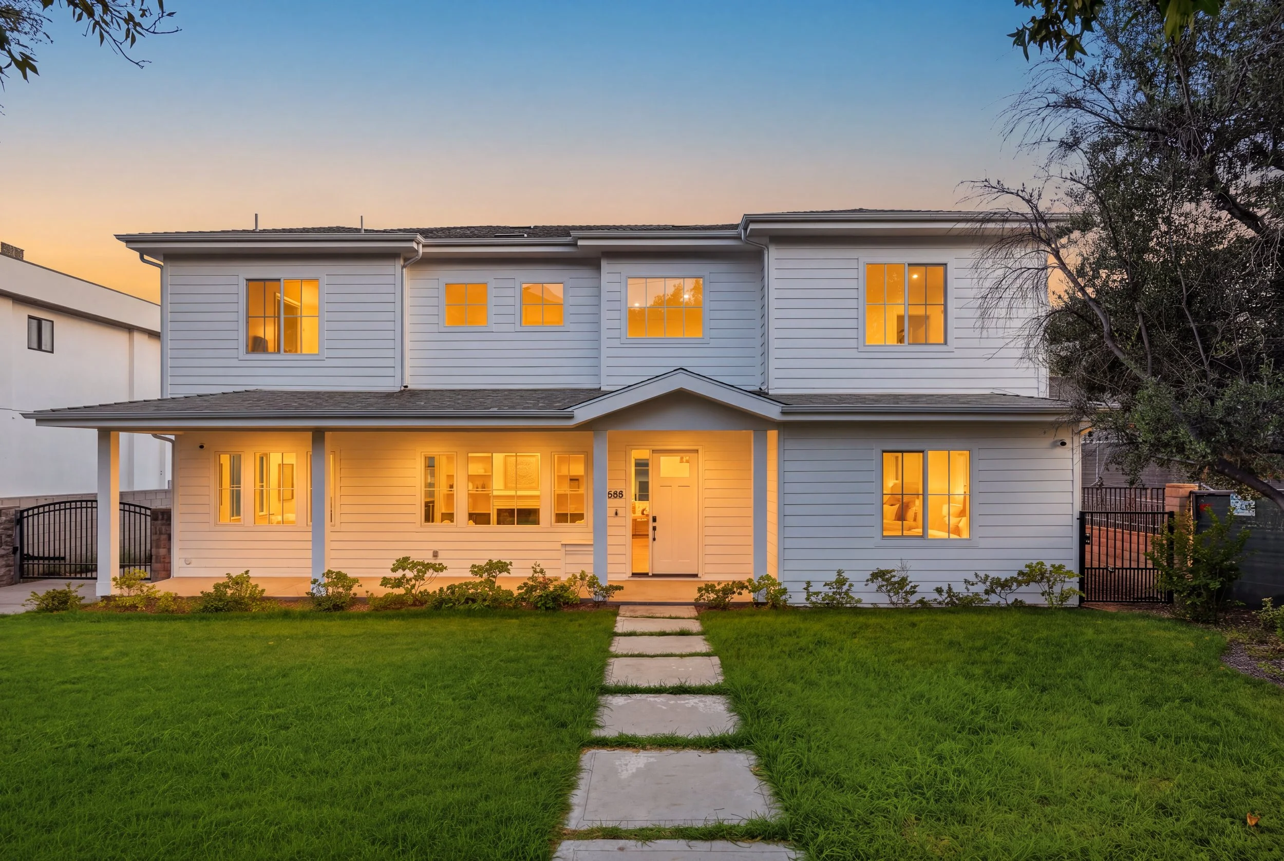 Front view of a white, two-story house at dusk with glowing interior lights, a front yard with a concrete pathway, green grass, and some plants. There are trees on the sides and neighboring houses visible.