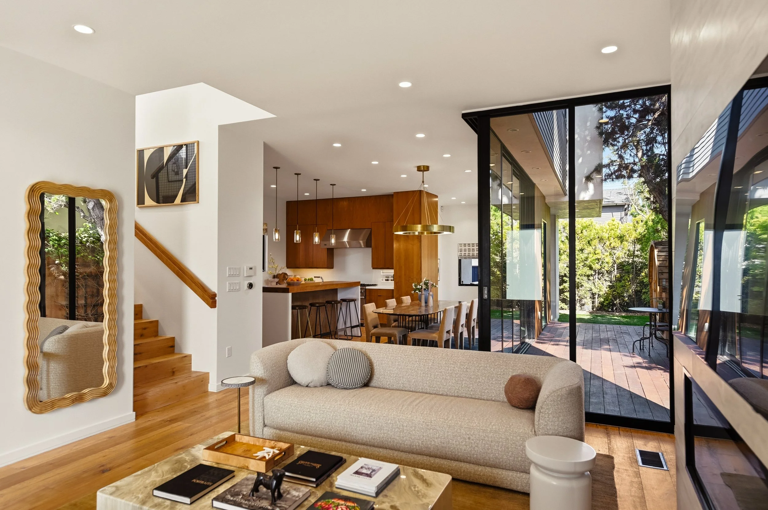 Interior of a modern open-concept living and dining area with large glass doors opening to a lush garden. A beige couch with pillows is in the foreground, a marble coffee table with books and decorations is nearby. The dining area features a wooden t