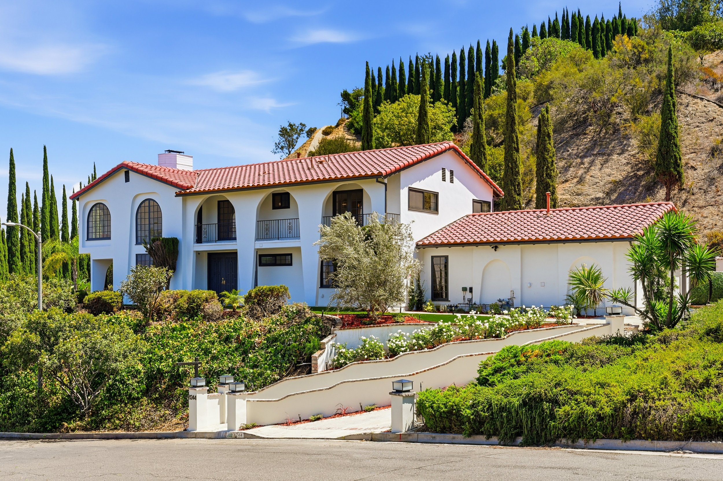 White house with red-tile roof on a hillside, surrounded by lush greenery and tall cypress trees, under a clear blue sky.