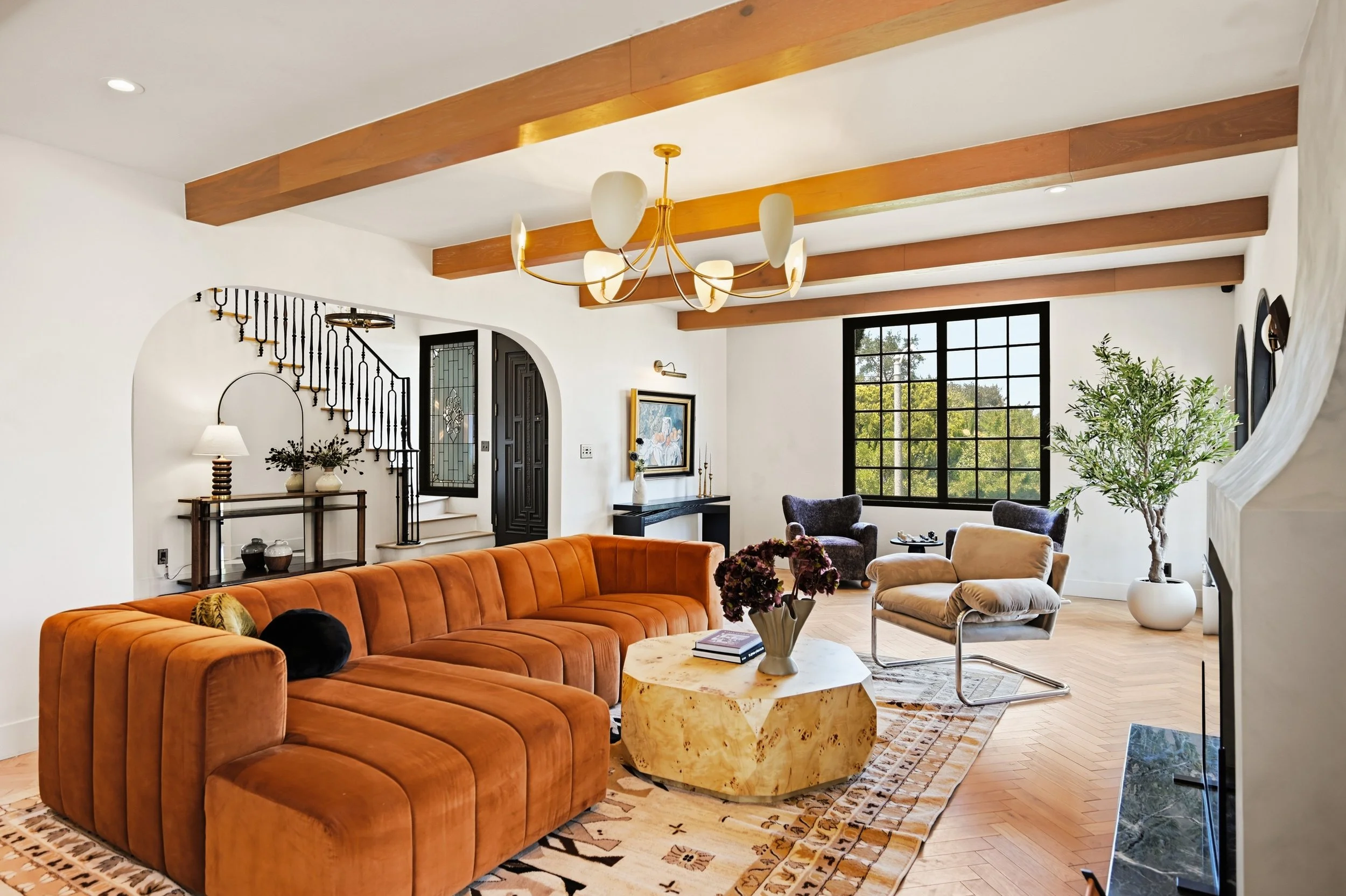 Living room with orange sofa, beige armchair, black chairs, large window, wooden beams on ceiling, chandelier, and indoor plants.