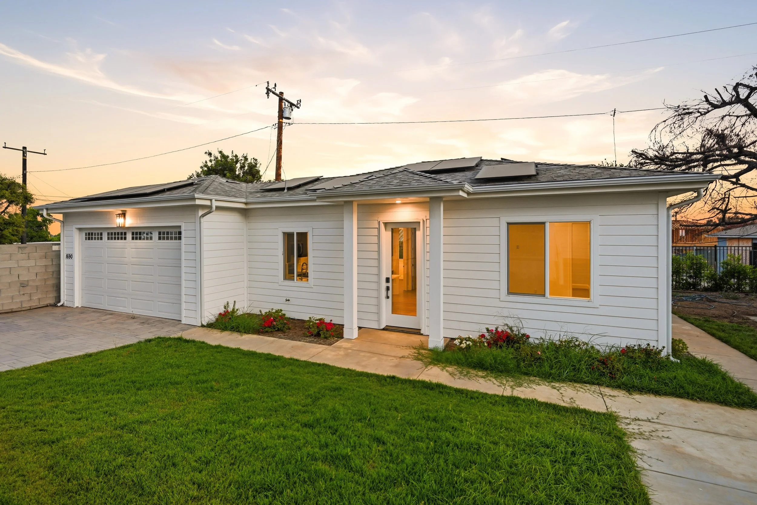 A white house with a garage door, front door, and windows, surrounded by a green lawn and garden with flowers, during sunset with a partly cloudy sky and solar panels on the roof.