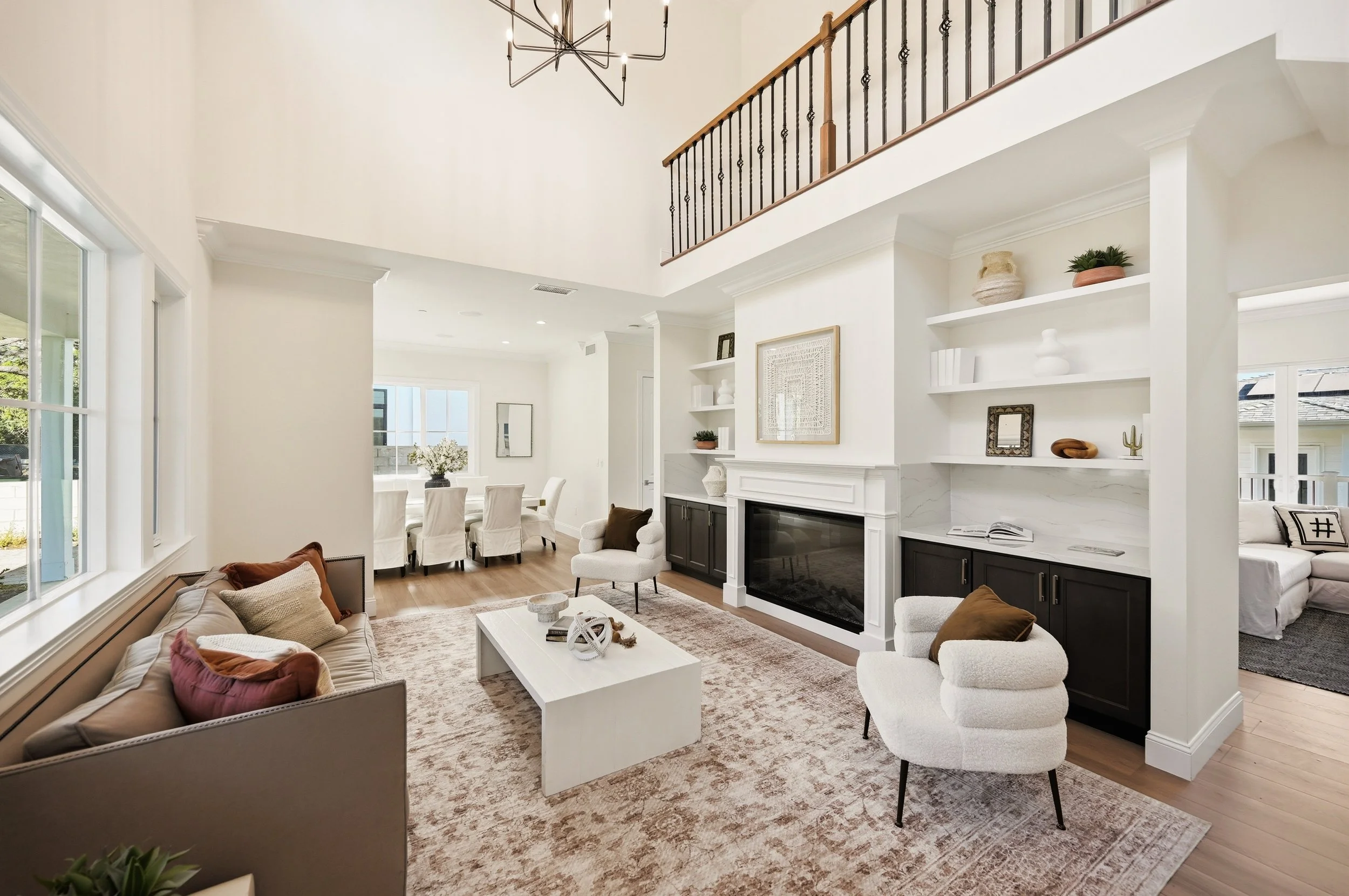 Interior of a modern living room with a high ceiling, large windows, a dining area with a white table and chairs, and a sitting area with a sofa, armchairs, and a fireplace, decorated with neutral and earthy tones.