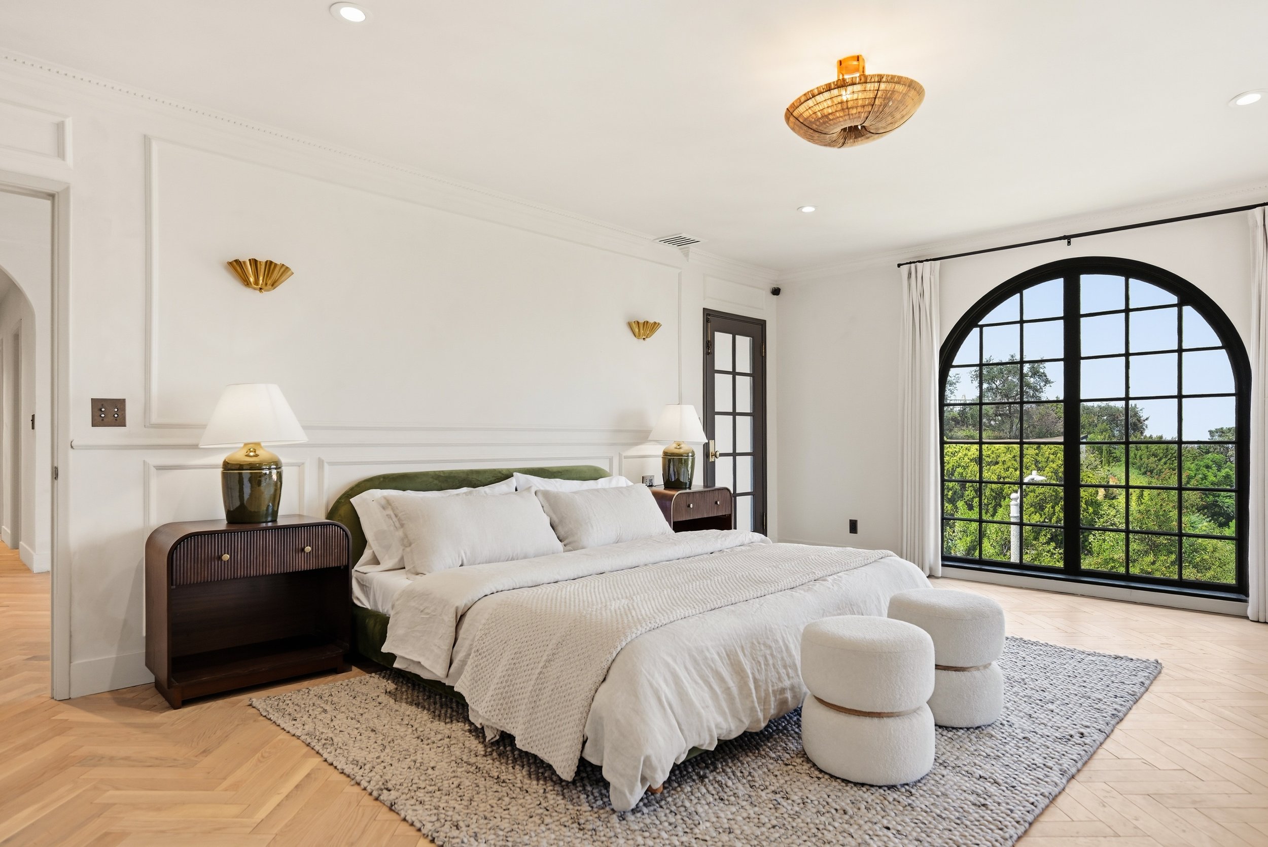 A bedroom with a large arched window showing green trees outside. The room has a white bed with a dark wooden headboard, two bedside tables with matching lamps, and decorative wall sconces. The floor is wooden with a textured area rug, and the ceilin