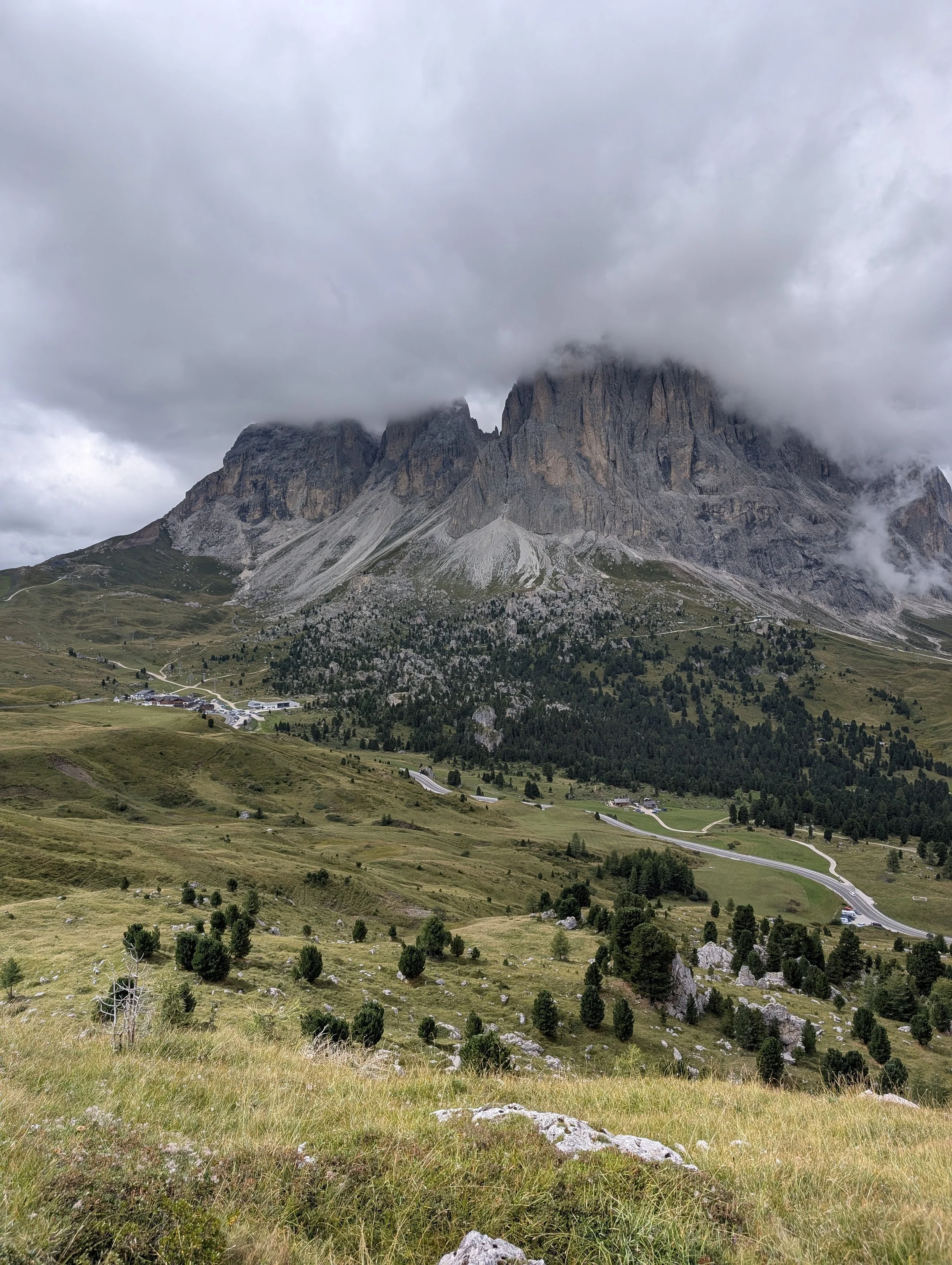 Scenic view of a mountain valley with tall rocky peaks partly covered by clouds, grassy meadows, scattered trees, and a winding road.