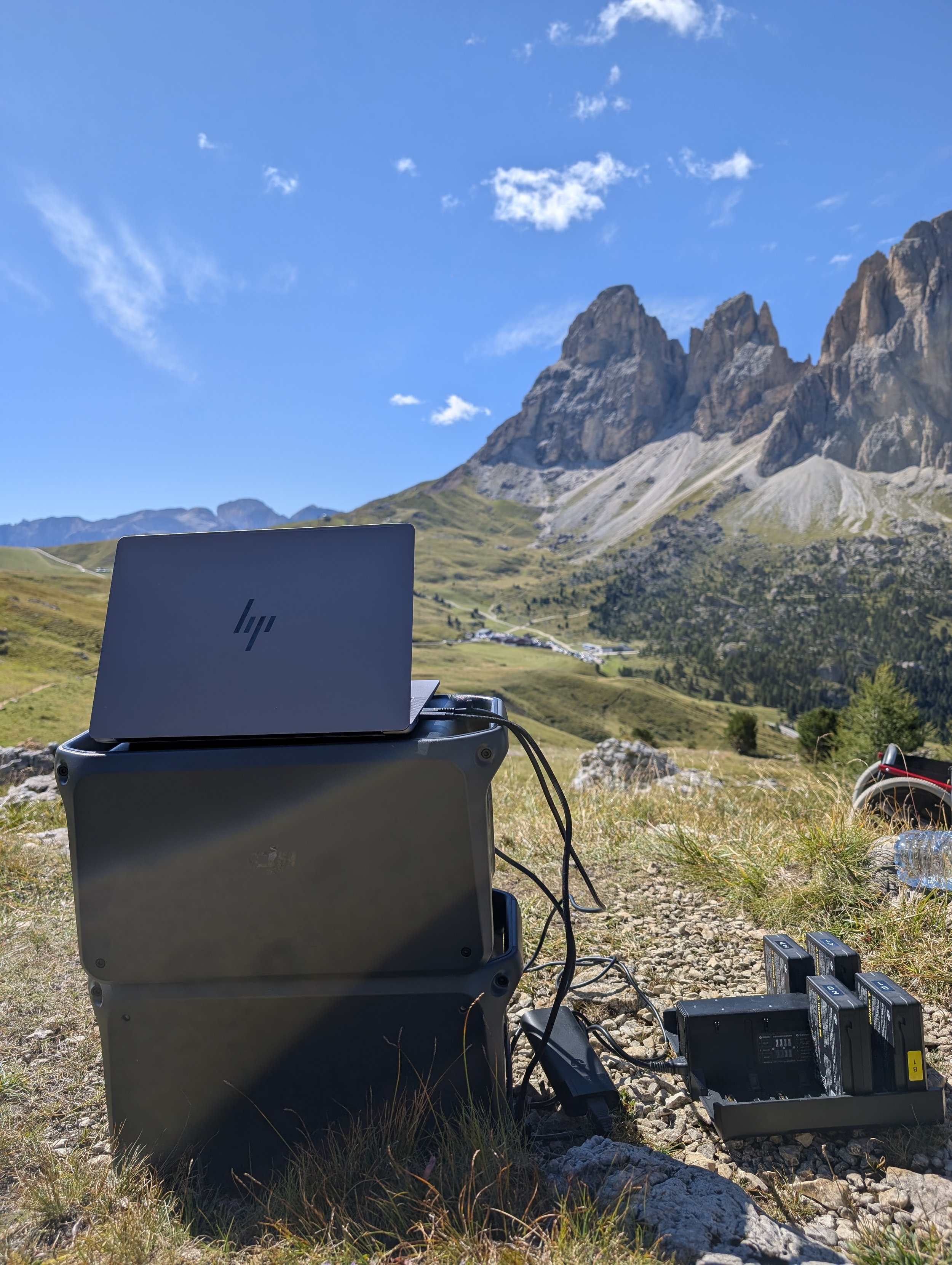 A laptop computer connected to electronic equipment outdoors in a mountainous area with rocky terrain, grassy patches, and a clear blue sky with a few clouds.