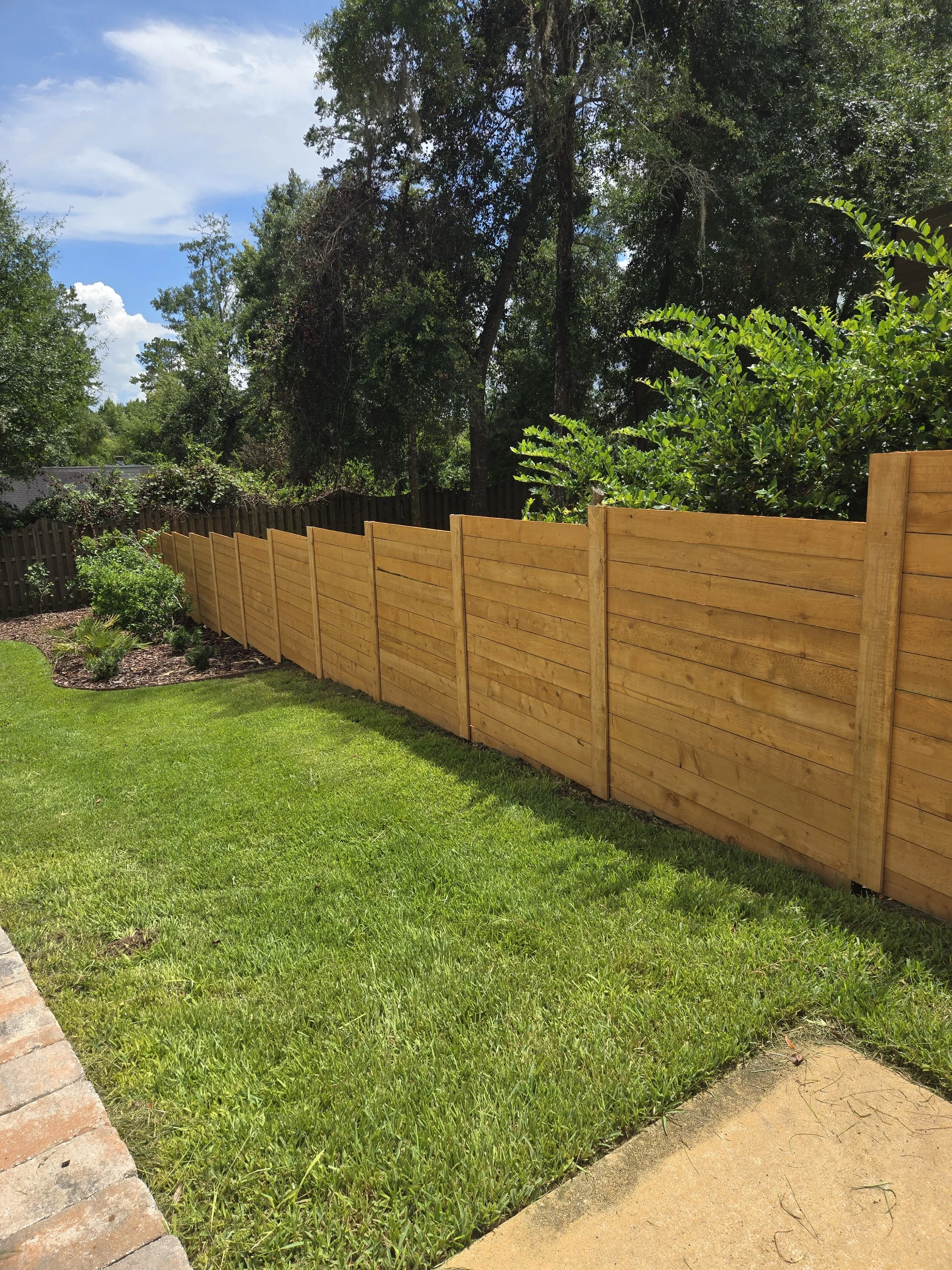 A backyard with a newly installed wooden privacy fence, green grass, and trees in the background under a partly cloudy sky.