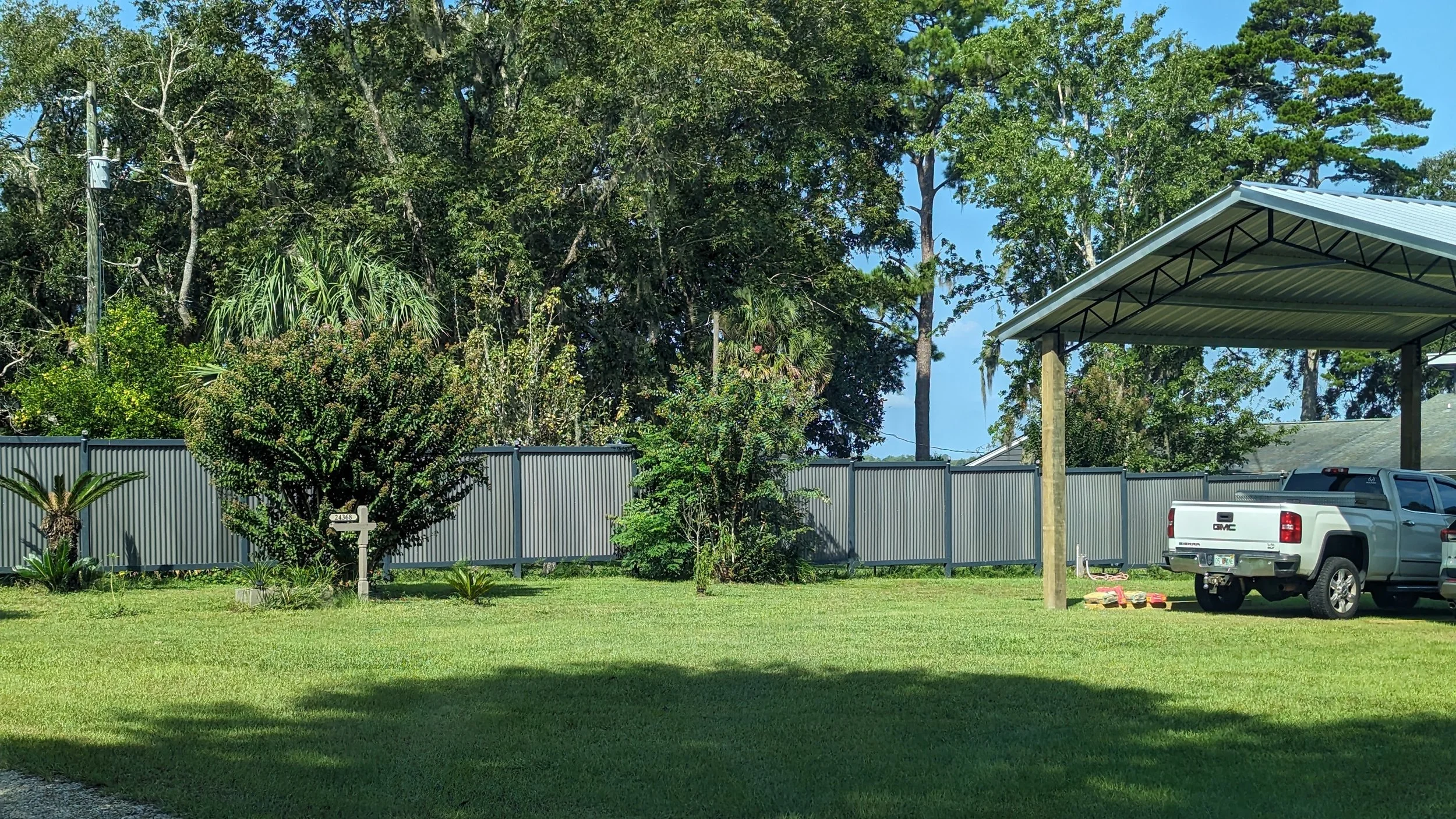 Residential backyard with green grass, trees, a gray fence, a white GMC pickup truck parked under a metal carport, and some gardening supplies on the ground.