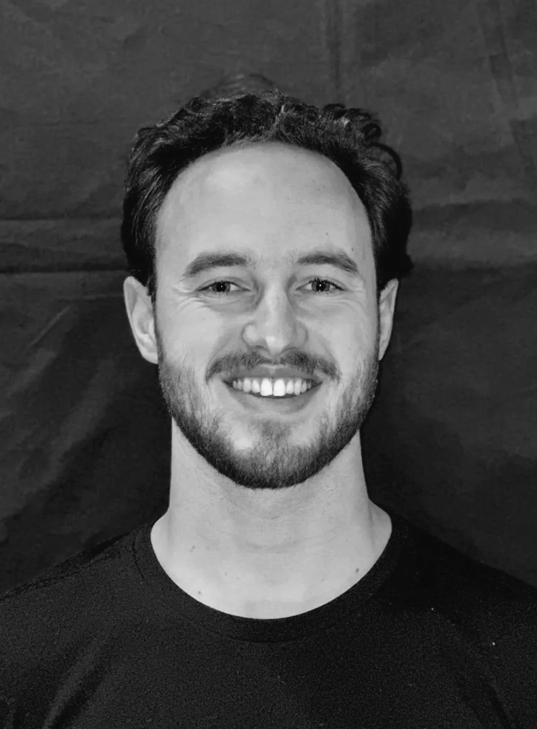 Black and white portrait of a smiling man with a beard, dark hair, and a black shirt, lying on a wooden surface.