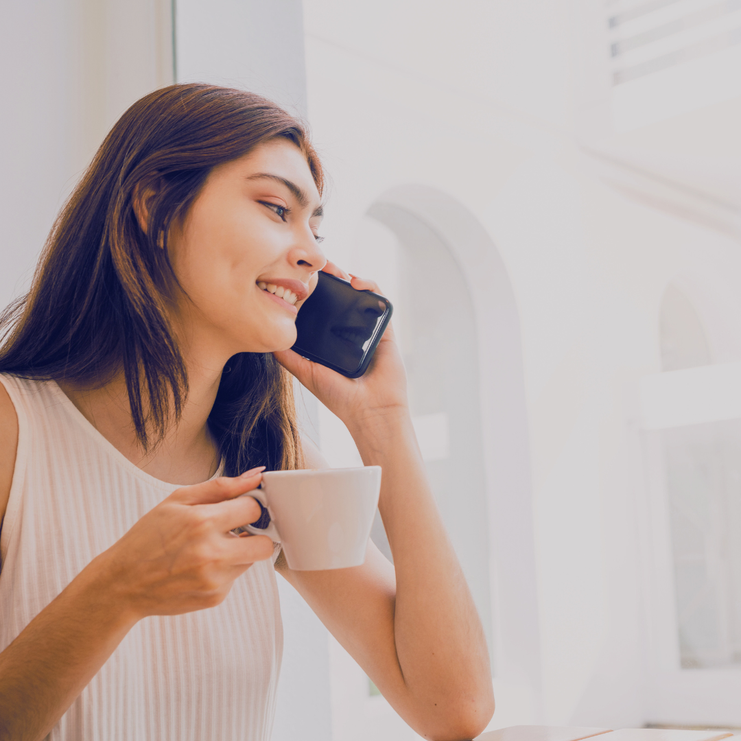 Woman smiling and talking on her cellphone while holding a coffee cup indoors near a window.