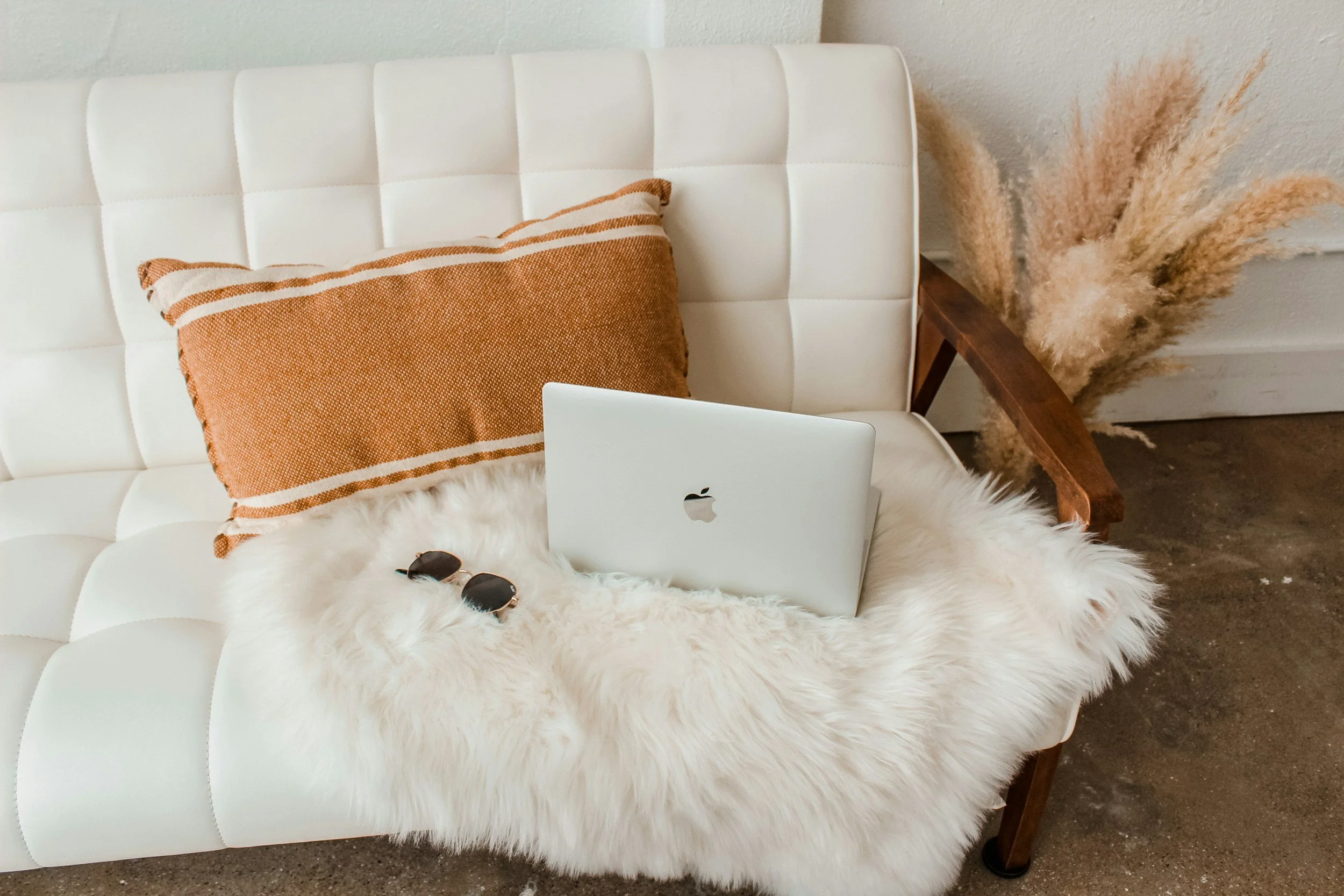 A cozy living room corner with a white tufted sofa, a brown decorative pillow, a white fluffy rug, a pair of sunglasses, a closed MacBook laptop, and a large vase with pampas grass.