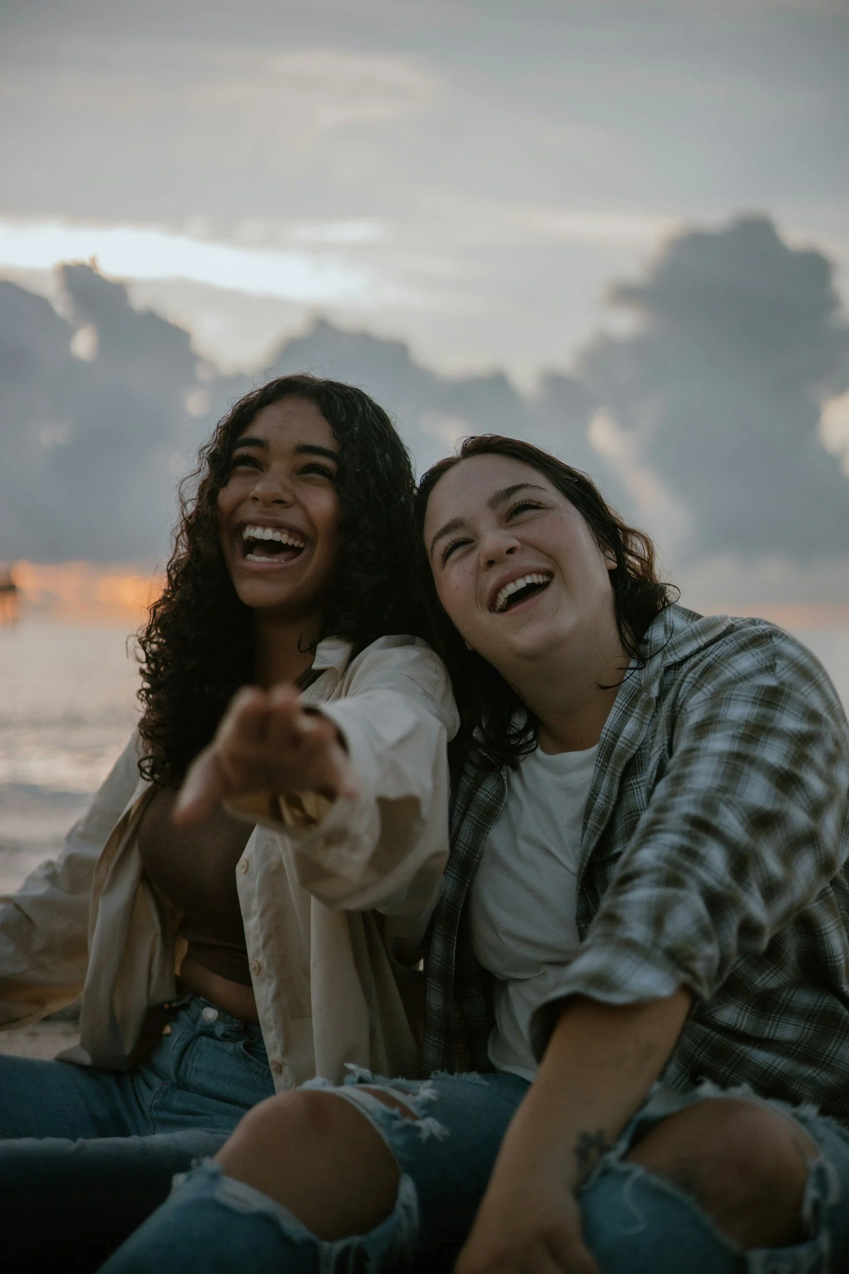 Two young women sitting on the beach during sunset, laughing and smiling, with clouds in the sky.