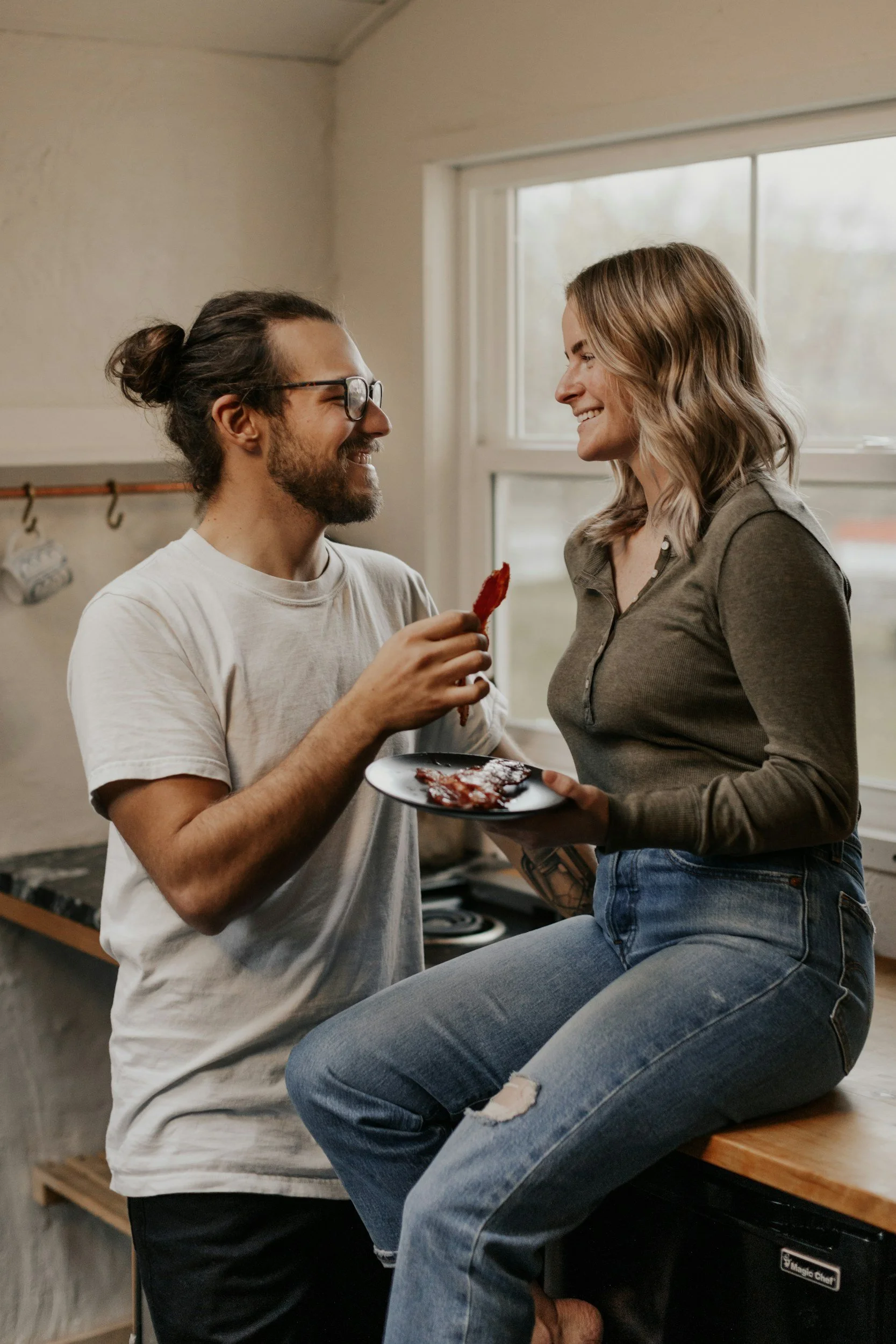 A man with glasses and a man bun holding a plate with bacon and interacting with a woman sitting on a kitchen counter, both smiling at each other.